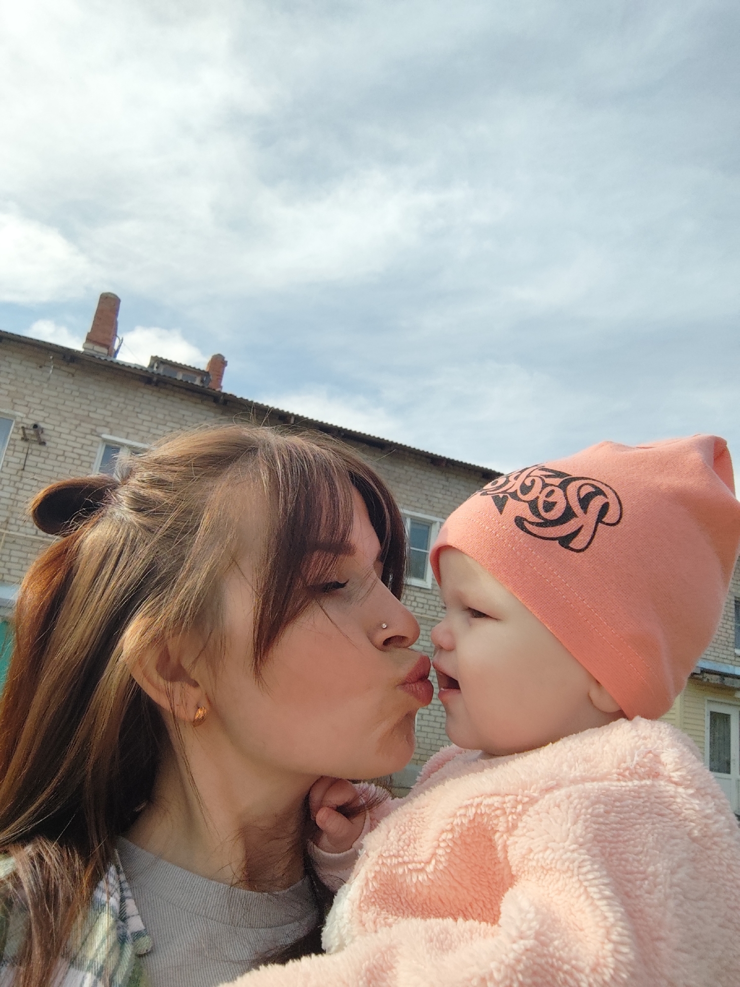 A medium shot captures a woman and a baby close to one another. The woman, with fair skin, has her brown hair pulled back with some strands framing her face. She wears a stud nose piercing and a pair of gold earrings. She wears a green and white plaid shirt over a gray shirt. She is puckering her lips as if to kiss the baby, who is dressed in a pink fuzzy outfit and a matching hat with a black design. The baby is looking at the woman. The photo is taken outdoors in daylight. A brick building is behind them, and the cloudy sky is in the background. The location is Фоки, Russia.