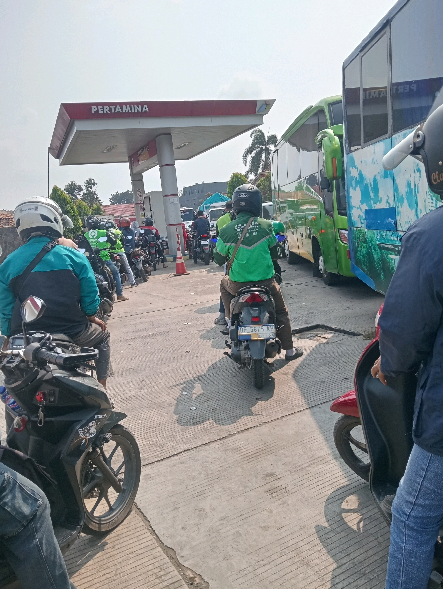 The image depicts a line of motorcycles and buses at a Pertamina gas station in Bandar Lampung, Indonesia. A large green bus with a scenic mural on its side is parked on the right, partially obscuring a smaller green bus. In front of the buses, several motorcycles are lined up, waiting for fuel. Some riders are wearing helmets and jackets, with one rider in a green jacket with the "Grab" logo, indicating he is a delivery driver. The gas station has a red and white canopy with the Pertamina logo. The ground is paved concrete, and the sky is clear and bright. The overall scene suggests a busy, ordinary day at a public service location.