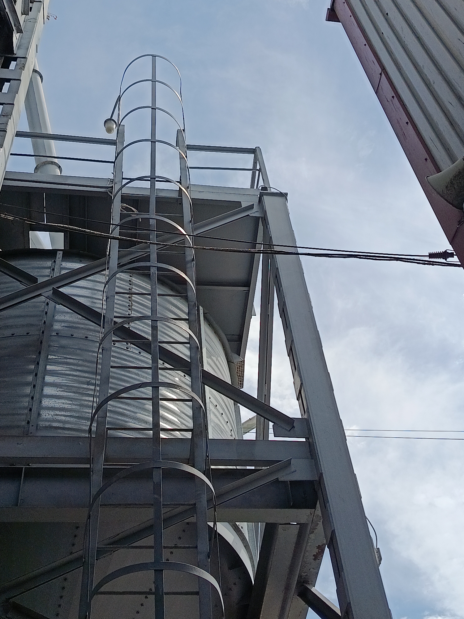 An exterior low-angle shot captures industrial silos under a cloudy sky in Biloziria, Ukraine. The metallic silos are supported by a grey steel structure, with a ladder featuring a protective cage leading up one of the silos. Overhead, power lines cross the frame, and a portion of another building with a red and grey facade is visible on the right. The lighting suggests it is daytime.