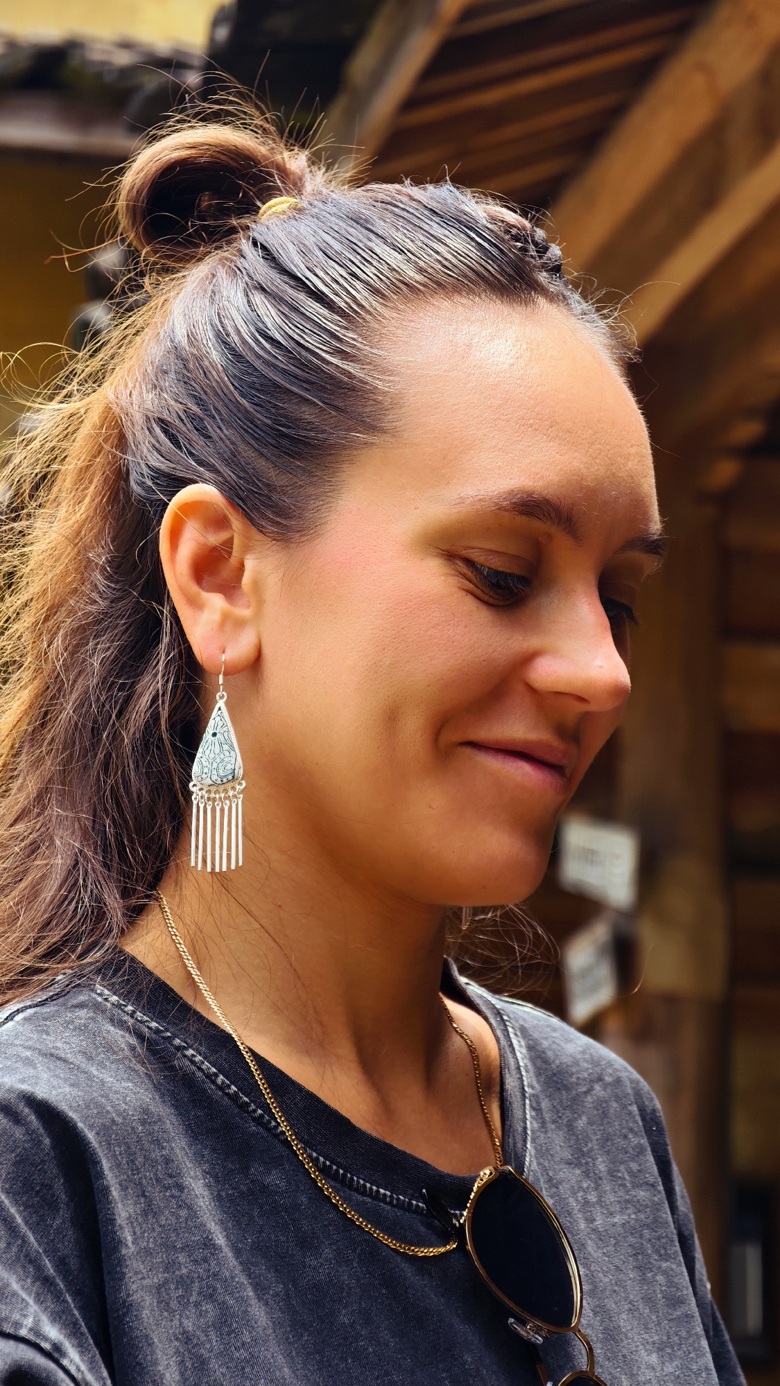 A woman with brown hair in a high bun wears a gray t-shirt, a gold necklace, and silver earrings in an outdoor setting. Sunglasses hang from the necklace. The background is blurred, but suggests a wooden structure. The image suggests a casual, perhaps tourist setting, although the location, Lũng Cú Commune, Vietnam, isn't visibly apparent in the scene.