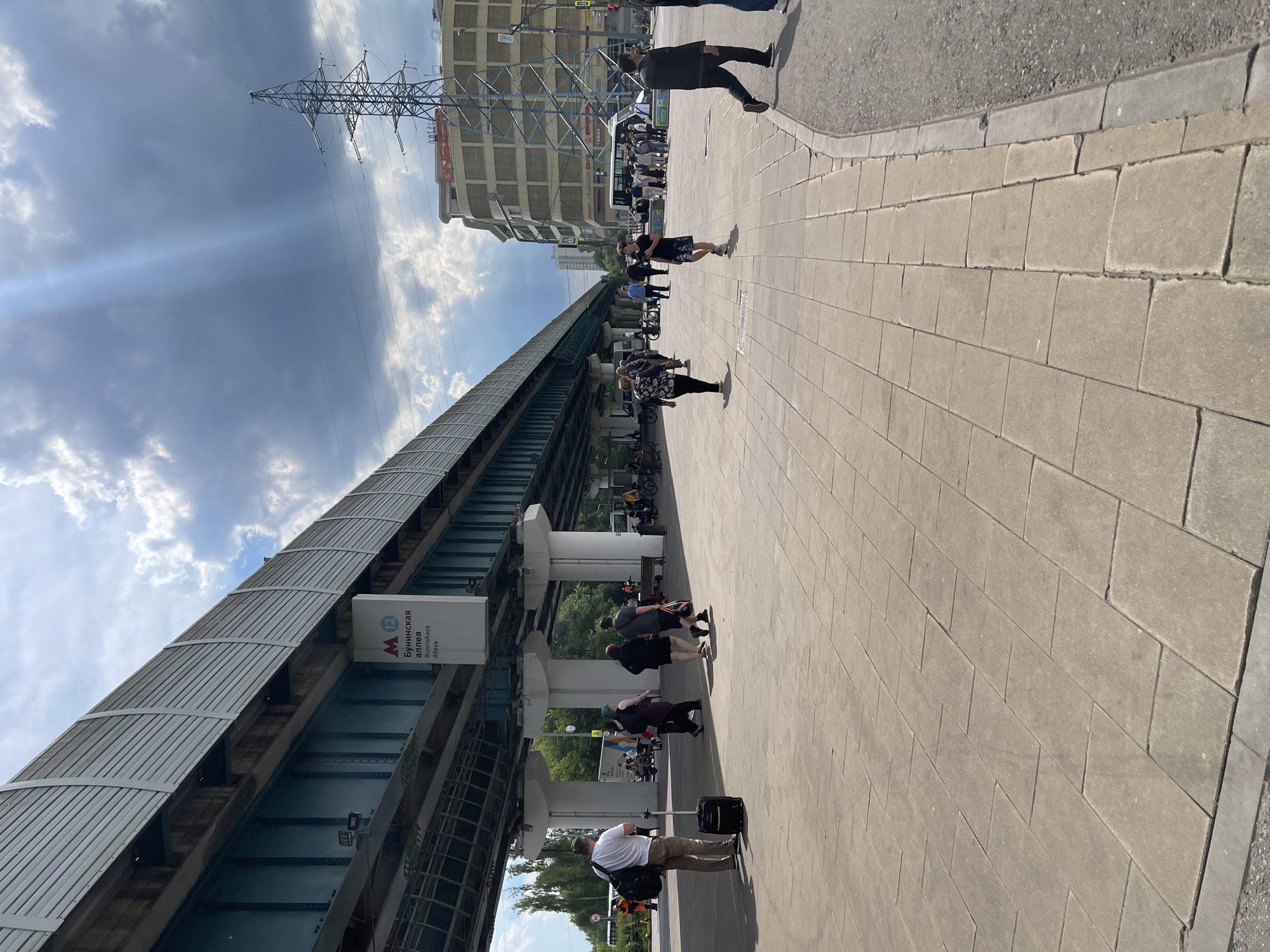 This vertical shot captures an urban scene in Moscow, Russia, featuring an elevated metro line, a sidewalk bustling with people, and buildings under a partly cloudy sky.
In the foreground, a light-colored paved sidewalk stretches from the bottom left towards the upper right. Several pedestrians are scattered across it. One man in a white t-shirt and tan pants stands with a black rolling suitcase and a shoulder bag in the immediate foreground. Other individuals and small groups walk or stand further along the sidewalk, some under the shade of the elevated metro structure.
The dominant feature on the left is the elevated track of a metro line, which has a distinct gray, ridged metal roof. A white sign attached to the metro structure clearly displays "М 12 Бунинская аллея" (Buninskaya Alleya) with the English transliteration "Buninskaya Alleya" below it, indicating a Moscow Metro station.
In the background, on the right side of the image, stands a large, multi-story building with a light-colored facade. Prominently visible on its exterior is a large red sign with the text "ОКЕЙ" (OKEY), likely a hypermarket. Other smaller signs on the building include "TABAK" (Tobacco) and "АКТИВ" (AKTIV). Behind this building, a tall, metal lattice power line tower is silhouetted against the sky.
The weather appears to be bright and clear, indicative of daytime, possibly late morning or early afternoon, with patches of blue sky and white clouds. Sunbeams are visible breaking through the clouds in the upper left portion of the sky. The overall scene depicts a typical busy urban environment with public transportation infrastructure.