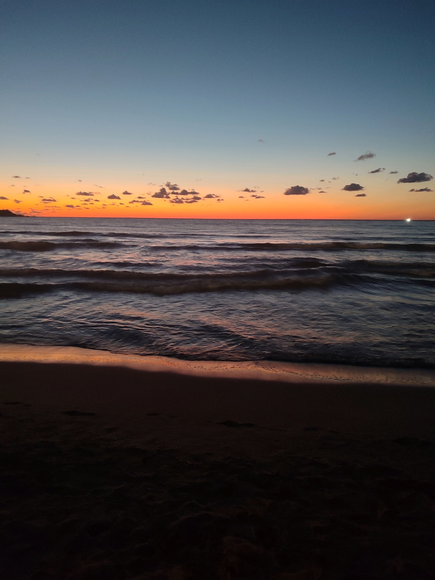 This is a low-angle, vertical, eye-level shot of a beach at sunset in Kandıra, Turkey. The sky transitions from a clear, deep blue at the top to a vibrant orange and yellow near the horizon, with scattered, small, dark clouds. The ocean is dark and choppy, with waves rolling towards the shore. The wet sand at the water's edge reflects the colors of the sky. The foreground is a dark, sandy beach, with footprints visible. The scene is devoid of people.