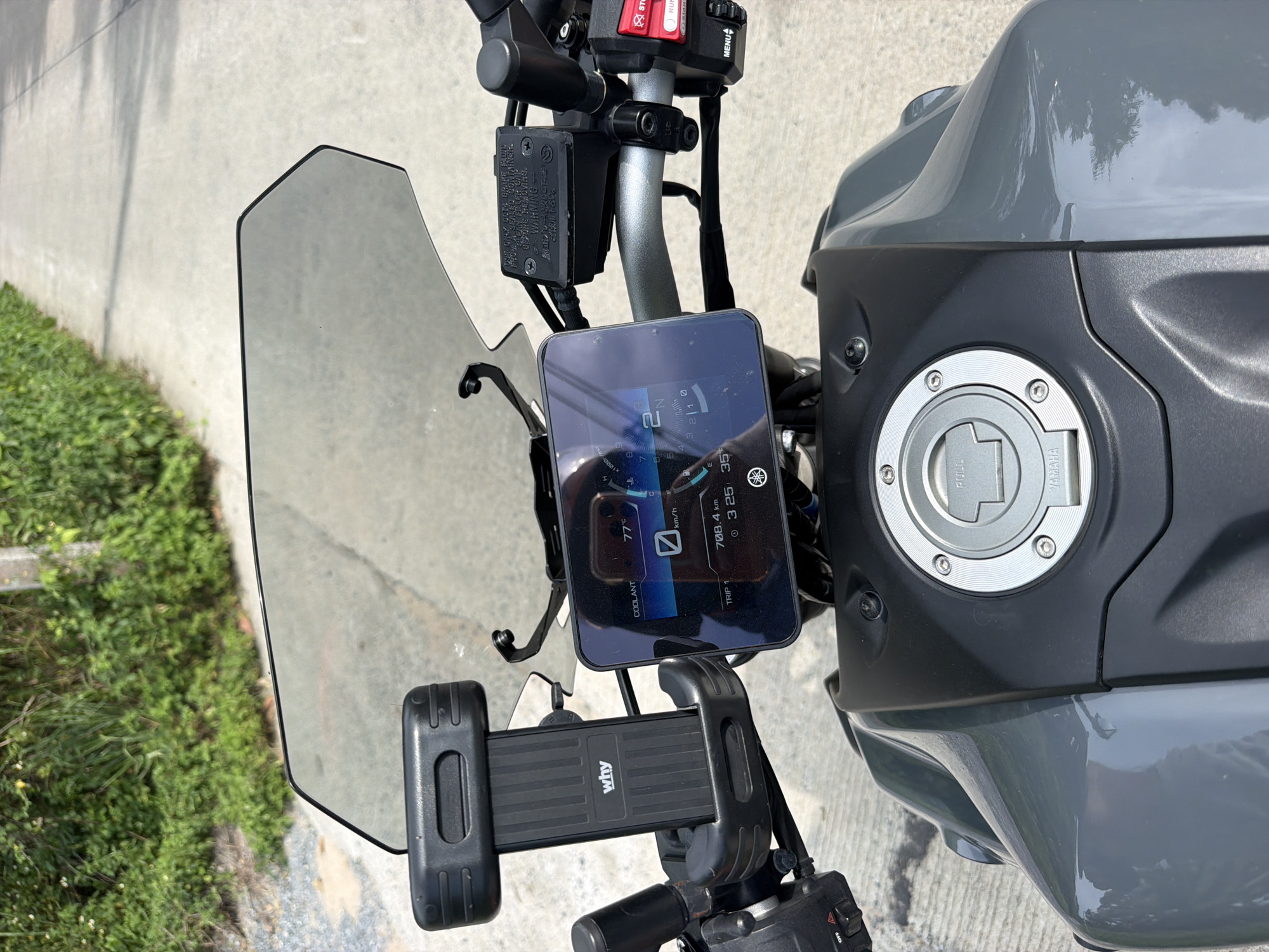 This is a first-person perspective photograph taken from a motorcycle parked on a paved road. The motorcycle's dashboard is the primary focus, displaying a digital screen with various information including temperature (77°C), speed (km/h), and trip distance (708.4 km) and time (3:25). A phone mount with the brand "why" is attached to the left handlebar. The motorcycle has a grey fuel tank with a silver cap. The background shows lush green vegetation on the left and a paved road extending into the distance. The scene appears to be set in a rural or suburban area, possibly Baan Mae Nam, Thailand, given the tropical foliage. The lighting suggests it is daytime with sunlight. There are no people visible in the image.