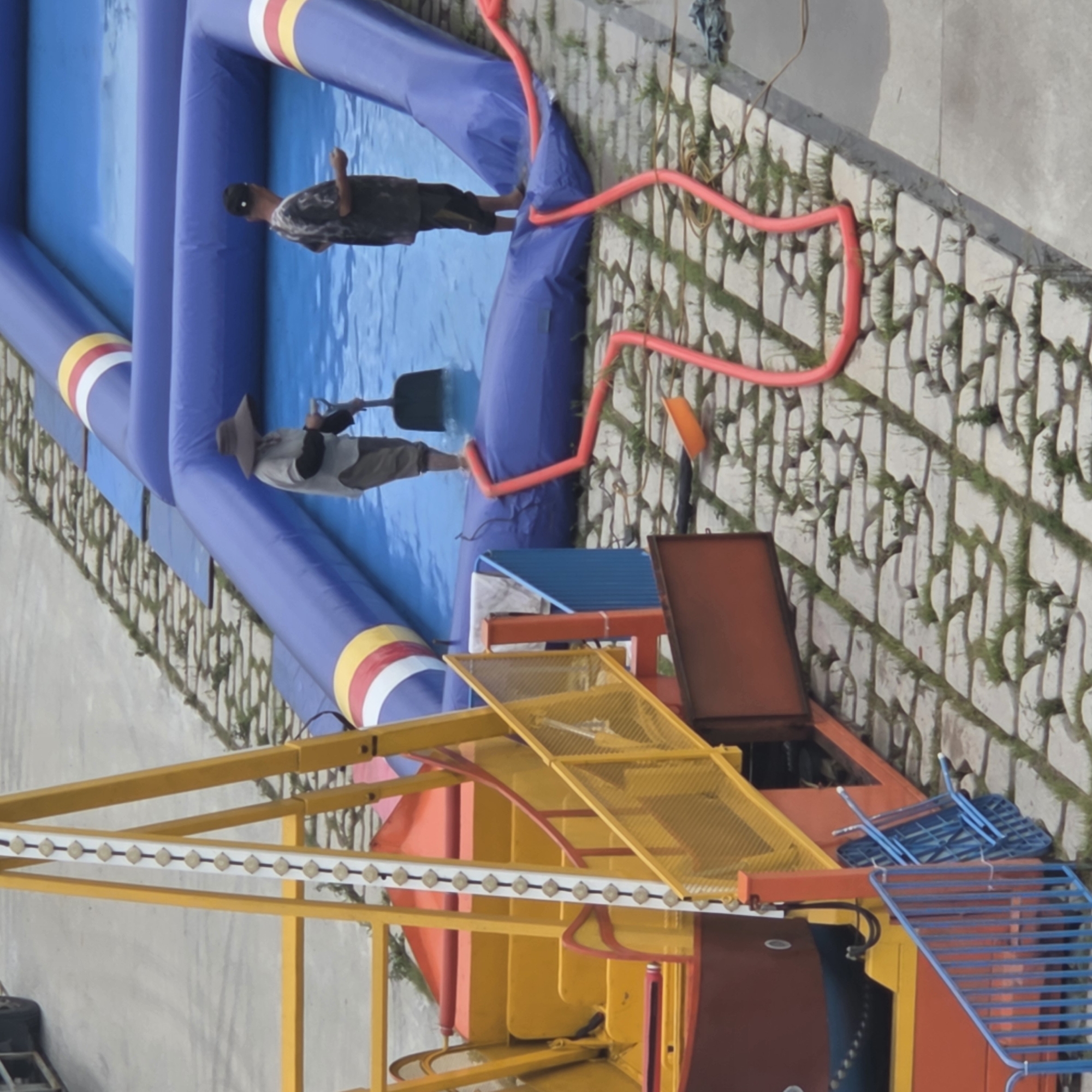 In Dunnae-myeon, South Korea, an aerial view captures an inflatable pool setup next to a carnival ride. Two individuals are present. One person, wearing a wide-brimmed hat and a light-colored shirt with dark sleeves, is standing in the blue water of the pool, holding a dark shovel. The other person, wearing a dark, patterned shirt and dark pants, is also in the water. The inflatable pool features blue tubing with red, yellow, and white striped accents. A bright red hose snakes across the paved ground, leading into the pool. To the left, a section of a yellow and red carnival ride with a Ferris wheel-like structure is visible. The ground is paved with interlocking rectangular stones, with patches of green grass growing between them.