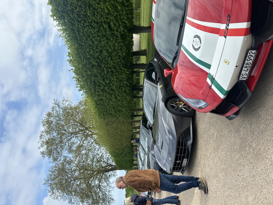 The scene takes place in the grounds of the Château de Chambord in the Loire Valley region of France, specifically in the landscaped gardens just west of the main chateau building. The image is captured from a ground-level perspective and showcases five individuals and four high-end sports cars.
Two individuals, a man and a woman, are standing in the foreground. To the left of the frame, a man in a brown jacket, blue jeans, and brown shoes stands with his back to the camera. He is engaged in a conversation with a woman in a dark jacket, her back turned to the camera. She is wearing blue jeans and black shoes.
There are two additional individuals visible in the background, also looking in the direction of the vehicles. They are positioned in the shade of the trees, their attire difficult to discern.
There are four sports cars present, all of the Ferrari brand. Two are positioned in the center of the frame. The closest to the camera is a red and white Ferrari with its hood up, showcasing a prominent Ferrari badge and a license plate reading "VS-488-992". It's followed by a dark grey Ferrari, its hood closed and its license plate obscured. Behind these two, a third dark grey Ferrari is partially visible, with its headlights obscured.
The vehicles are parked along a paved path, which disappears into the background amongst the trees and manicured gardens. In the background, a row of neatly trimmed trees creates a barrier between the path and the surrounding grounds. The trees, with their vibrant green foliage, are situated against a backdrop of a bright, cloud-filled sky.
