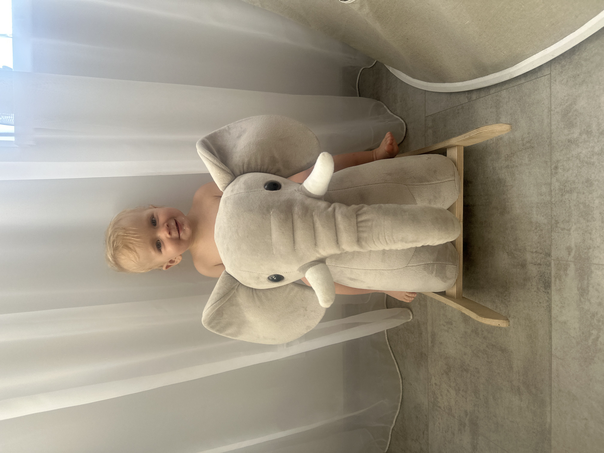 A small child with blonde hair and light skin is positioned on a beige plush elephant rocker. The child is looking at the camera. The scene takes place indoors, as indicated by the tiled floor and white sheer curtains in the background. The location is not discernable from the image.