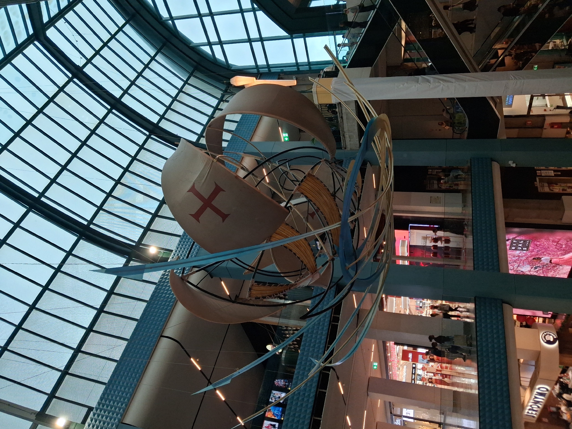 The image captures a large, abstract ship sculpture suspended within a shopping center environment, located in Lisbon, Portugal. The sculpture, resembling a historical sailing vessel, is constructed from various materials including sail-like forms with a red cross emblem. Below the sculpture are multiple levels of retail stores, recognizable by the presence of shop fronts and mannequins behind glass. The upper reaches of the image display a grid-patterned glass ceiling that floods the space with natural light. The people within the retail levels appear as indistinct figures due to the distance.