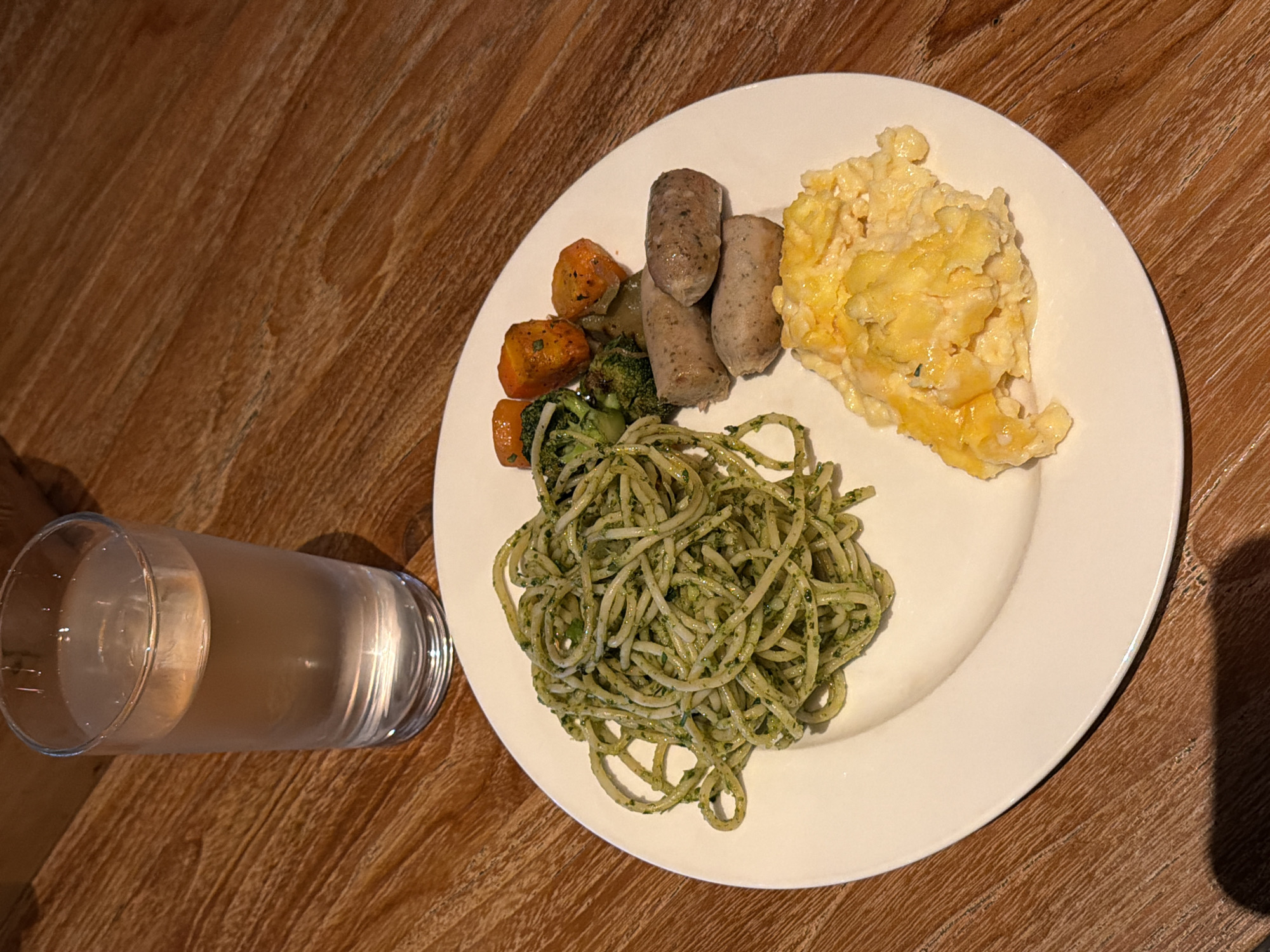 A close-up, eye-level, medium shot shows a white plate of food on a wooden table. The plate contains spaghetti with pesto, three sausages, scrambled eggs with cheese, and a side of steamed vegetables including broccoli and carrots. To the left of the plate, a clear glass filled with water and ice is visible. The lighting is natural and slightly warm, casting soft shadows. The image appears to be taken in a restaurant or dining setting. The wooden table has a visible grain and texture. The specific location in Kuta, Indonesia is not discernible from the image itself.