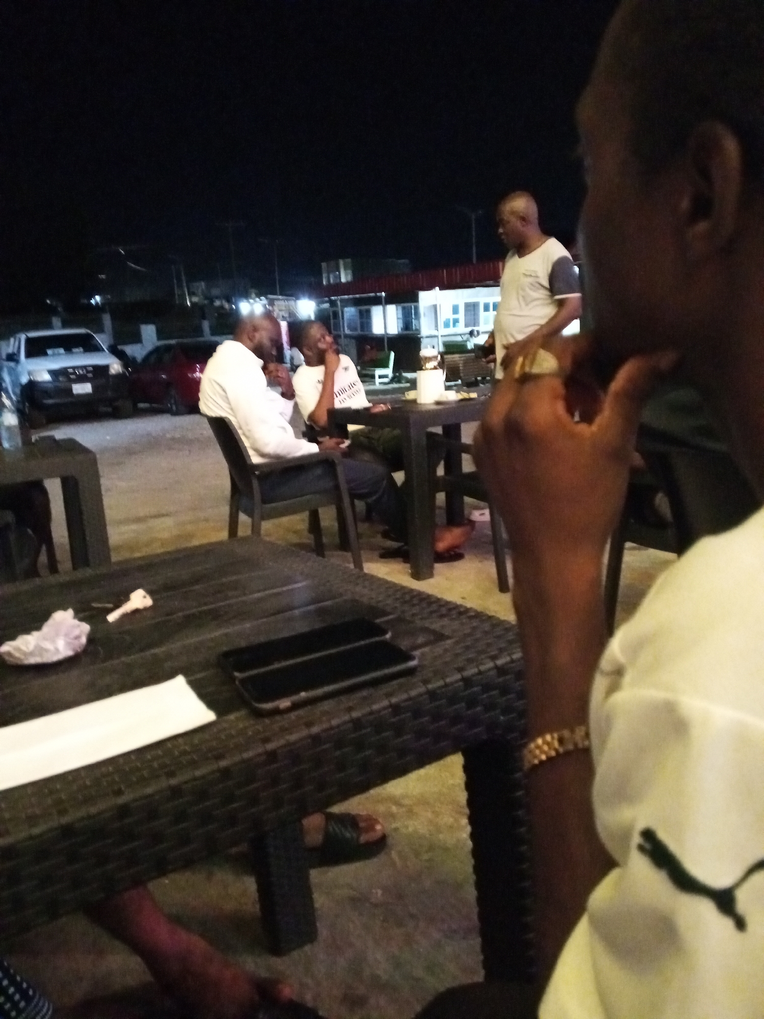 The image depicts an outdoor scene at night. Four individuals are visible. Two men are seated at a table engaged in conversation. One man stands near them, appearing to be addressing them. A fourth individual is in the foreground, partially visible, wearing a white shirt with a black Puma logo, and is resting their hand on their chin.
Various tables and chairs are scattered around the area. On the table in the foreground are two mobile phones, a white paper or napkin, and a crumpled paper. In the background, vehicles, including a white pickup truck, and a building with a red roof are visible.
Given the prompt, it is assumed this scene is set in Lokoja, Nigeria. The exact location within Lokoja cannot be determined without more specific information.