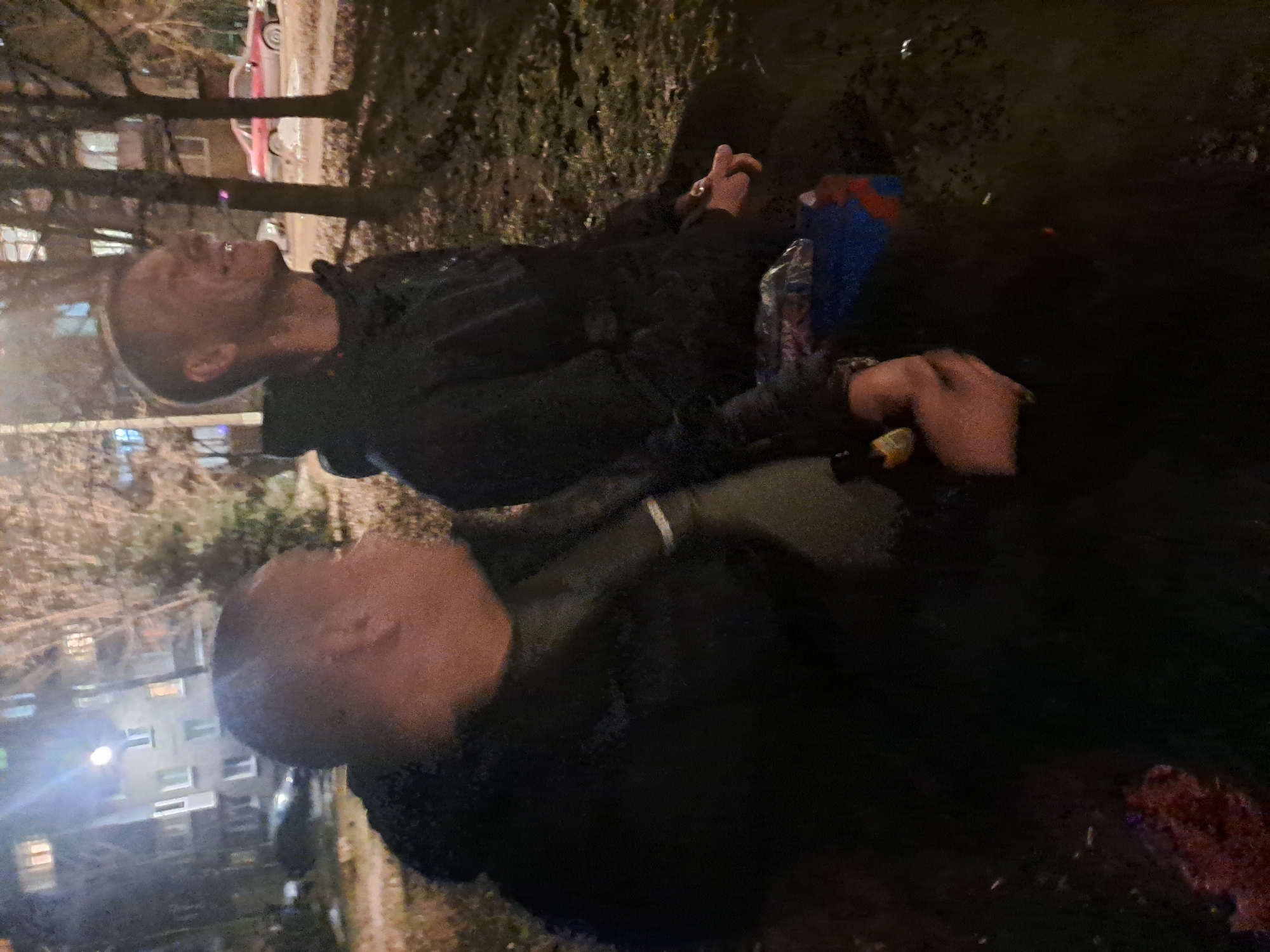 Two men are lying on the ground outdoors, possibly in a park or near a residential area. The setting appears to be in the evening or at night. The men are wearing dark clothing. There are trees and buildings in the background. A car is visible in the background. The location is possibly Elektrostal, Russia, based on the query.