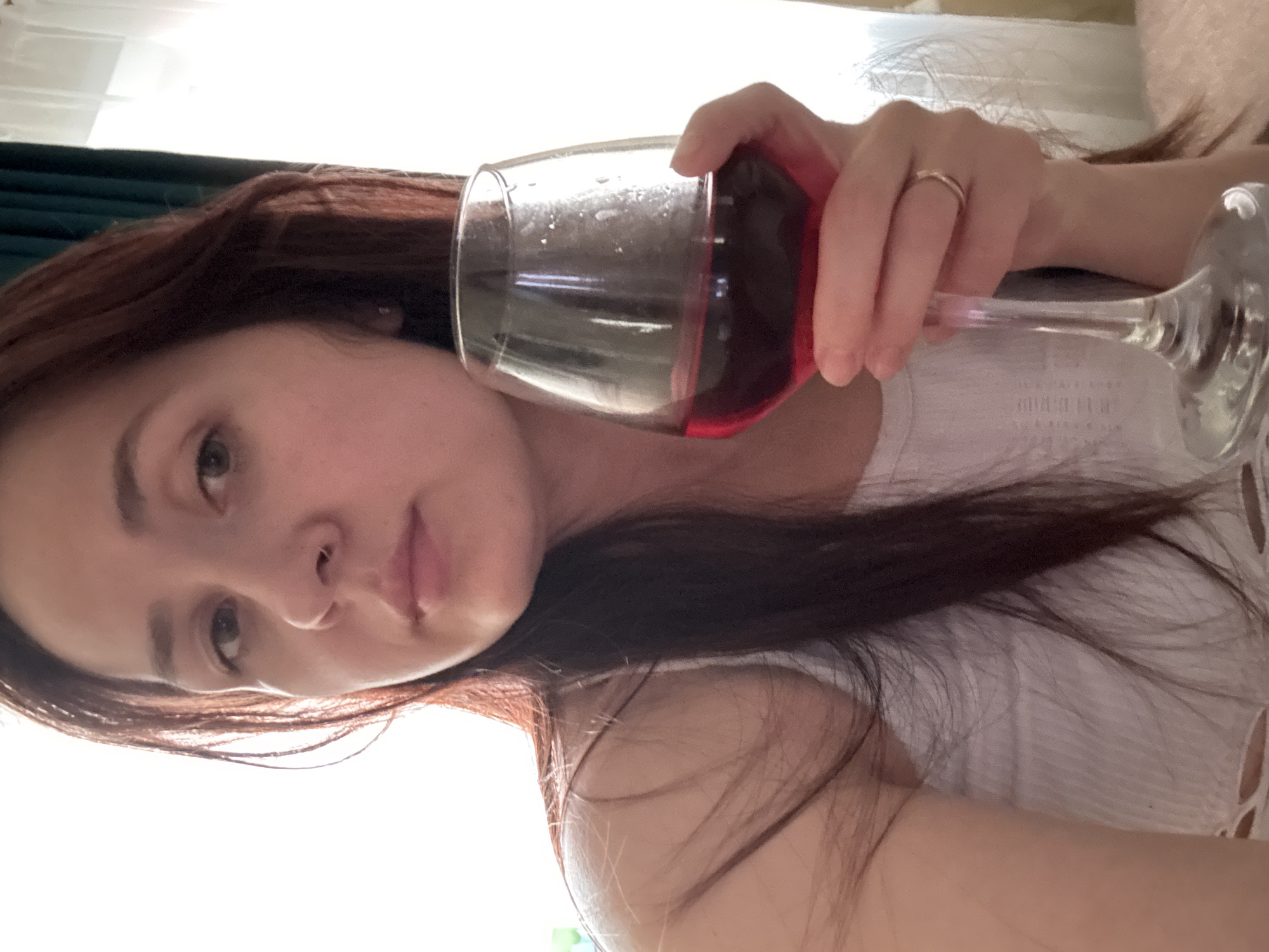 The image shows a selfie of a young woman with long, dark hair holding a stemmed glass of red wine. She is wearing a white tank top. Her face is partially illuminated by bright light coming from the left side of the frame, creating a high-contrast scene. She has a nose ring and is wearing a ring on her right hand, which is holding the wine glass. The background is indistinct, with a hint of curtains on the right. There is no discernible information in the image to indicate a specific location in the city of Naberezhnye Chelny, Russia.