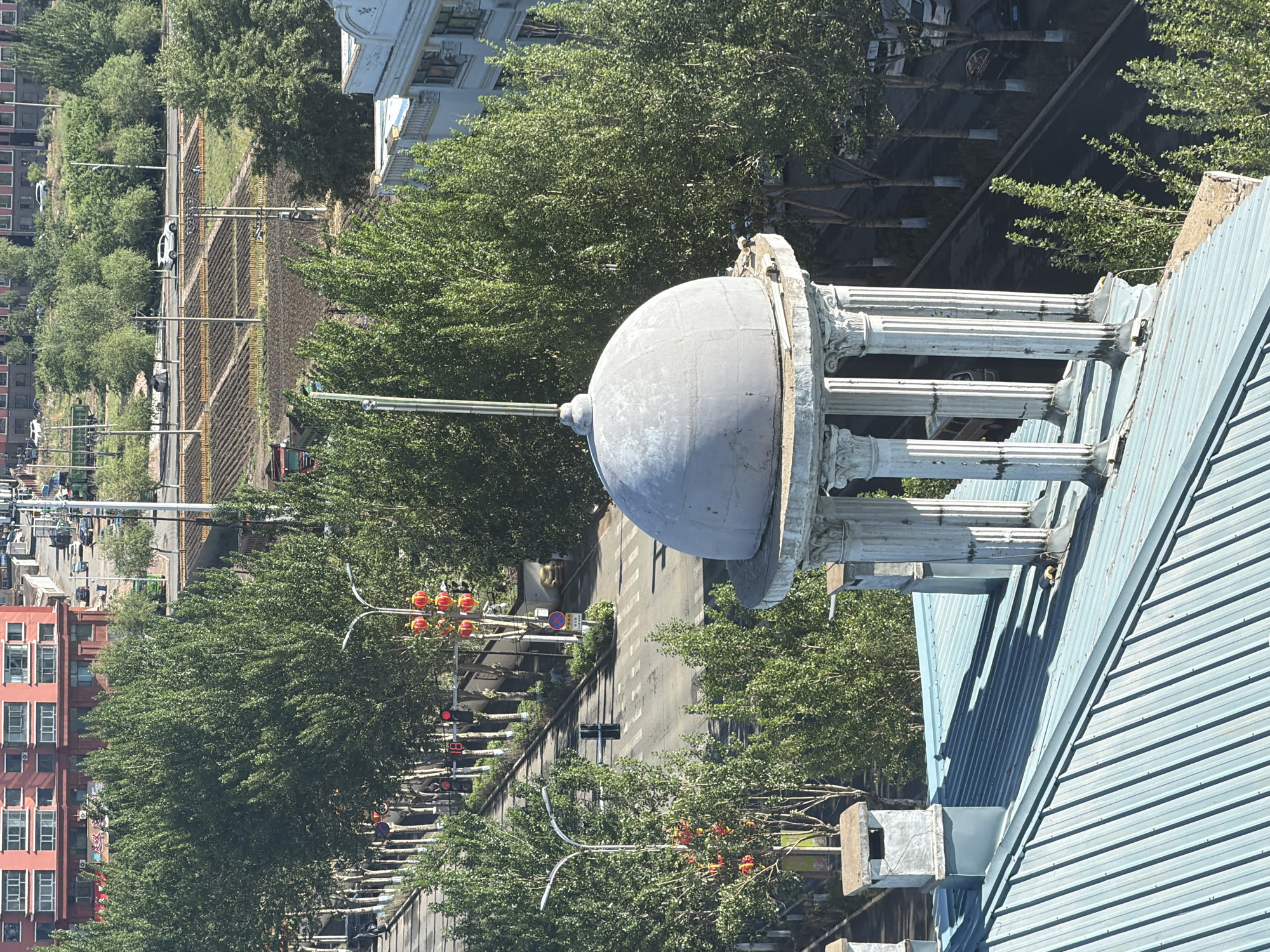 An elevated view captures a sunlit urban street in Tongjiang, China, bordered by buildings and abundant greenery. In the foreground, positioned on a light blue corrugated metal rooftop, stands a weathered, grey dome structure atop a circular colonnade featuring decorative pillars with Corinthian-style capitals. Nearby on the roof are square-shaped vents or chimneys.
The midground features a multi-lane city street with a tree-lined median strip. Several cars, predominantly white, are visible on the road, with some appearing to be parked. The street is flanked by numerous lush green trees with dense foliage, some bearing orange-red fruit or flowers. On the left side of the street, a multi-story building with a red-orange facade and numerous windows is visible, displaying air conditioning units beneath several windows. To the far upper right, part of a lighter-colored, ornate building is discernible.
Traffic infrastructure includes multiple streetlights and traffic signals. One red traffic light clearly displays the number "18," likely indicating a countdown. A "no parking" sign (red circle with a diagonal line over a 'P') is also visible near the street. The bright daylight and strong shadows suggest a clear, sunny day, likely mid-morning or afternoon. No specific event is depicted beyond general city traffic and activity. The overall impression is a bustling urban environment with a mix of architectural styles and considerable greenery.