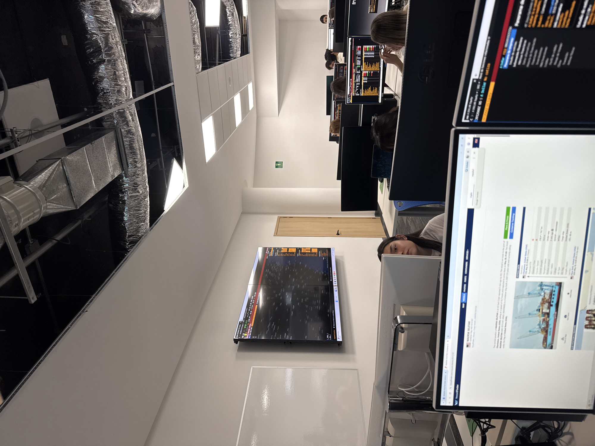 The image shows an office space in Puebla, Mexico. The office contains multiple computer workstations where people are working. Visible objects include monitors, keyboards, and office equipment. The office has exposed ductwork in the ceiling. A large monitor is mounted on the wall, displaying financial data. A whiteboard is also visible on the wall.