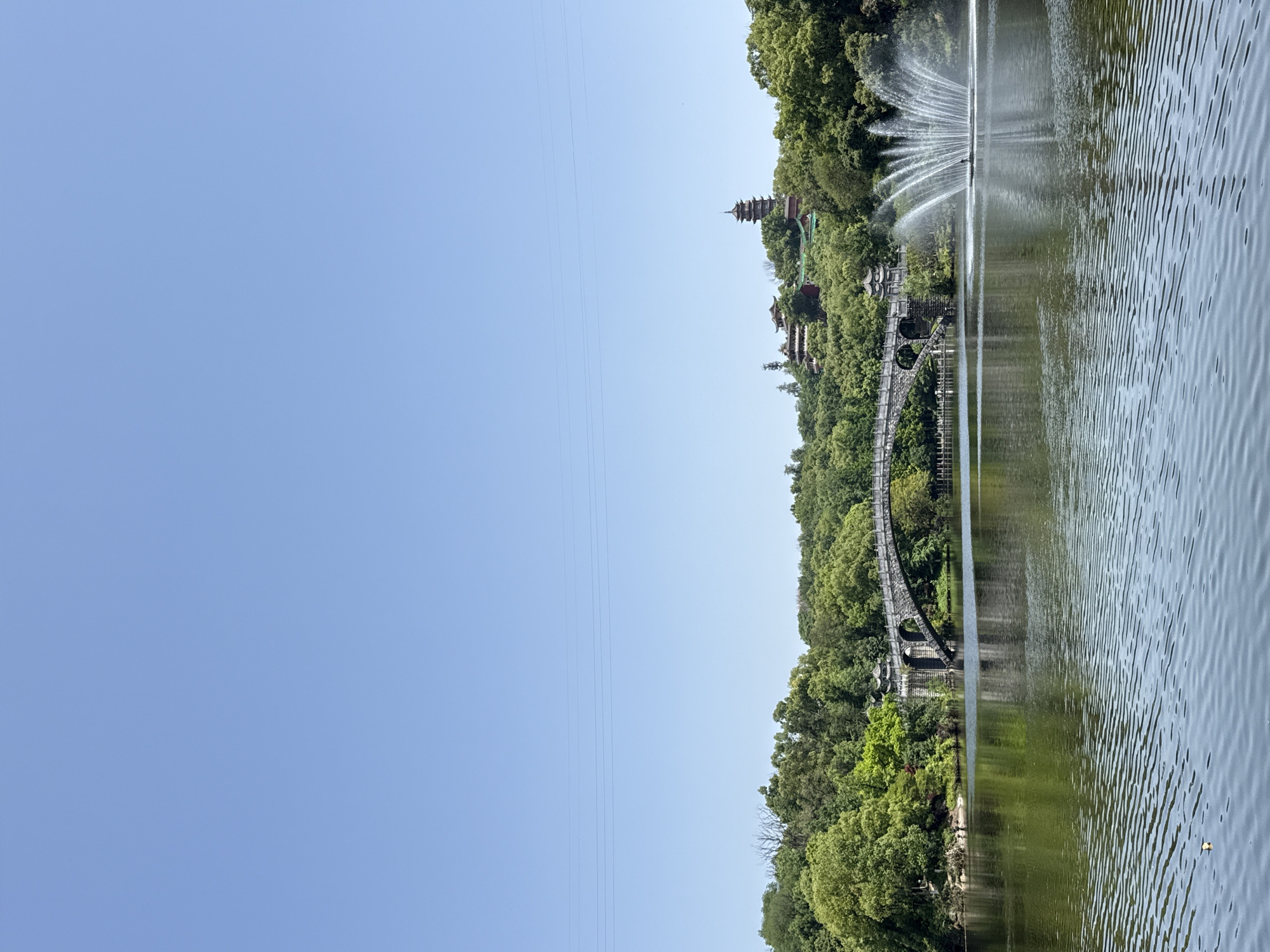 The image depicts a serene landscape in Jiulongpo District, China, featuring a body of water reflecting the surrounding environment. On the far bank, lush green trees cover the terrain, interspersed with architectural elements, including a traditional pagoda structure and a bridge. A fountain is centrally positioned in the water. Power lines are visible in the clear blue sky. The water surface displays gentle ripples, reflecting the surrounding greenery and sky.