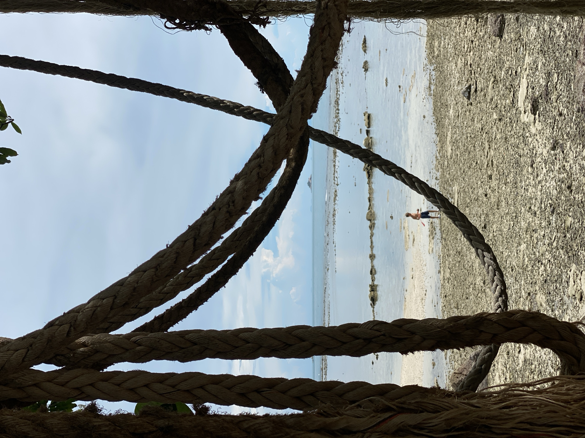 The image shows a beach scene in Koh Phangan, Thailand. In the foreground, thick ropes are draped across the frame, creating a layered effect. Beyond the ropes, a person is visible walking on a sandy and rocky shore. The water is calm and shallow, with a clear sky overhead, suggesting it is daytime with fair weather. The overall impression is one of a relaxed, natural environment.