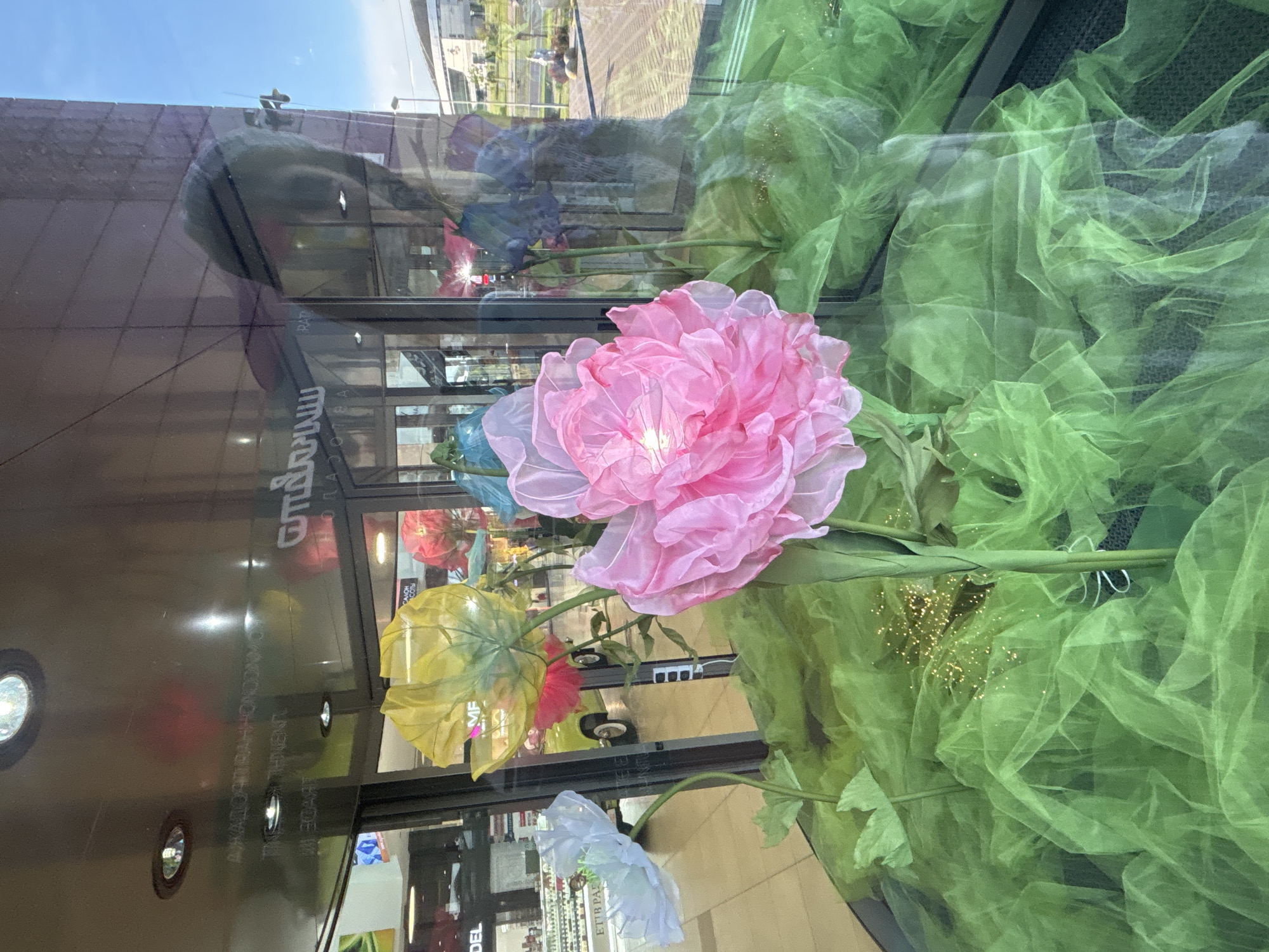 An upward-oriented photograph captures a vibrant display of large, artificial fabric flowers, viewed through a reflective glass surface. The main subject is a prominent pink flower, made of sheer, crinkled fabric, positioned centrally in the frame. Below it are yellow and white flowers of similar material, and glimpses of blue and red flowers are visible in the background and reflections. These flowers are set against a backdrop of voluminous, crinkled green tulle fabric, some of which has golden glitter sprinkled on it.
The glass surface creates significant reflections, revealing a bright blue sky and modern-looking buildings with glass facades and some with patterned exteriors, suggesting an urban or commercial setting. A street or road with what appears to be a yellow vehicle is also reflected. Inside the display space, round spotlights are visible in the reflections.
Visible text on the glass or reflected includes Russian words in Cyrillic, such as "ЖАЛОБА ОДНАЖДЫ" (A complaint once), "МИРОВЫЕ" (World), and "САЛОН СОФЬИ" (Sofia's Salon), along with partially readable English text like "UNREAL THADE IM" (possibly "UNREAL TRADE IN") and "ETTB PAVI", and "DEL". The bright reflections and clear sky indicate it is daytime. The overall scene suggests a shop window or an interior display in a commercial area, possibly a mall in Minsk, Belarus, given the context.