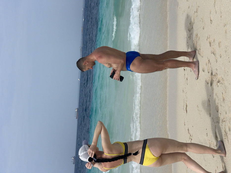 An outdoor scene depicts two individuals on a sandy beach in Zanzibar, Tanzania, facing the ocean. A female, positioned on the left, stands with her back to the camera, wearing a yellow two-piece bikini with thin black horizontal stripes on the top and bottom. She wears a white mesh baseball cap and has long dark hair styled in a single braid down her back. Her hands are raised, appearing to adjust her cap or hair. A male stands to her right, facing slightly left, shirtless, wearing blue speedo-style swim briefs with a white "Speedo" logo. He holds a black insulated tumbler or bottle with both hands, looking down at it. Both individuals are barefoot, though faint outlines suggest light-colored water shoes are present. The foreground consists of light-colored sand with visible footprints and small wave ripples. The ocean behind them displays a distinct transition from clear turquoise shallow water to darker blue deeper water further out. Small white-capped waves break near the shore. Several large cargo ships and at least one white-sailed vessel are visible on the distant horizon under an overcast or hazy light-blue sky.