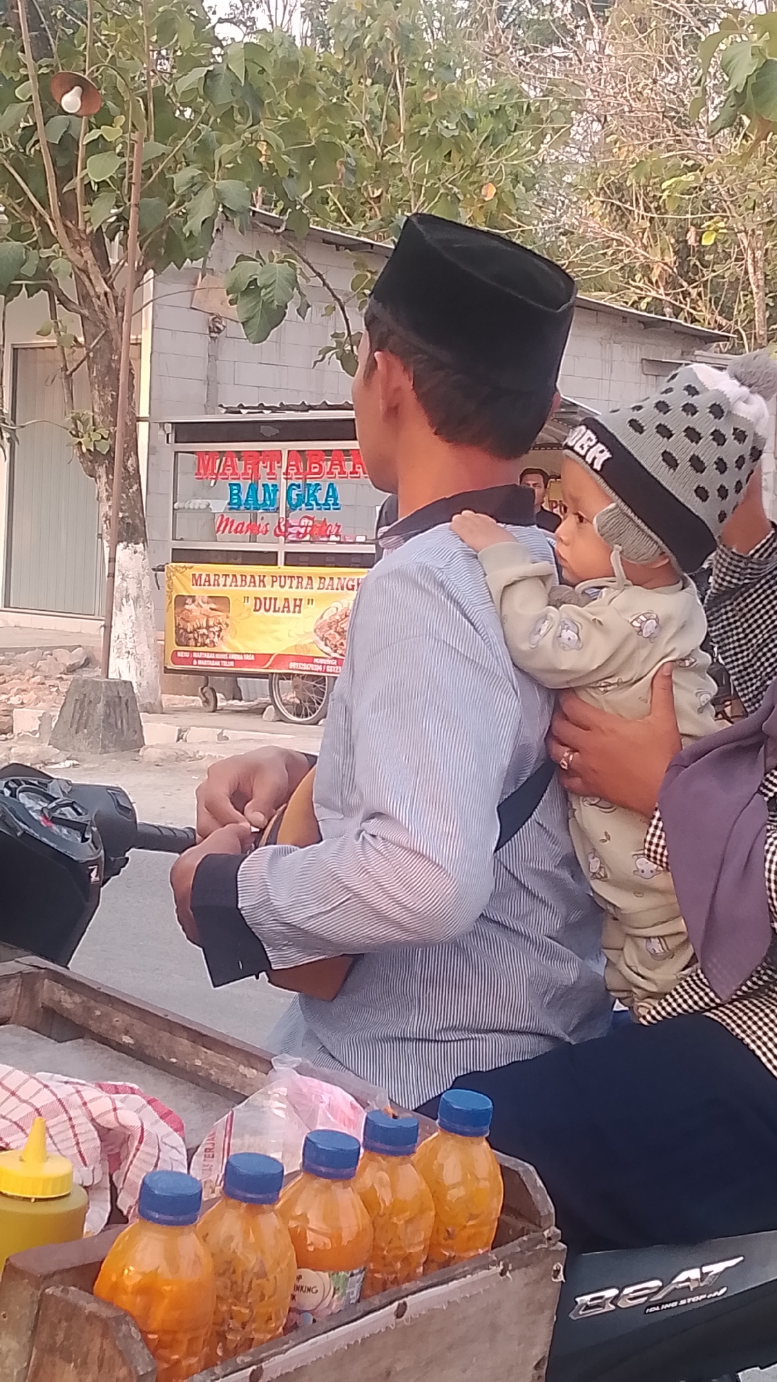In Karanglangu, Indonesia, a man wearing a black peci hat rides a motorcycle. He is carrying a baby on his back, who is wearing a grey and white patterned hat. The man is wearing a blue and white striped long-sleeved shirt. The baby is wearing a light-colored onesie with animal prints. To the right of the man and baby, another person is visible, wearing a purple hijab and a black and white checkered long-sleeved shirt. In the background, a food stall with yellow signage is present. The signage reads "MARTABAK BANGKA" and below that "MARTABAK PUTRA BANGKA 'DULAH'". In the foreground, a wooden cart is filled with several orange bottles with blue caps, likely beverages, and a red and white checkered cloth. The scene appears to be outdoors during the day, with trees and a building visible in the background. The motorcycle has a "BEAT" logo on its side.