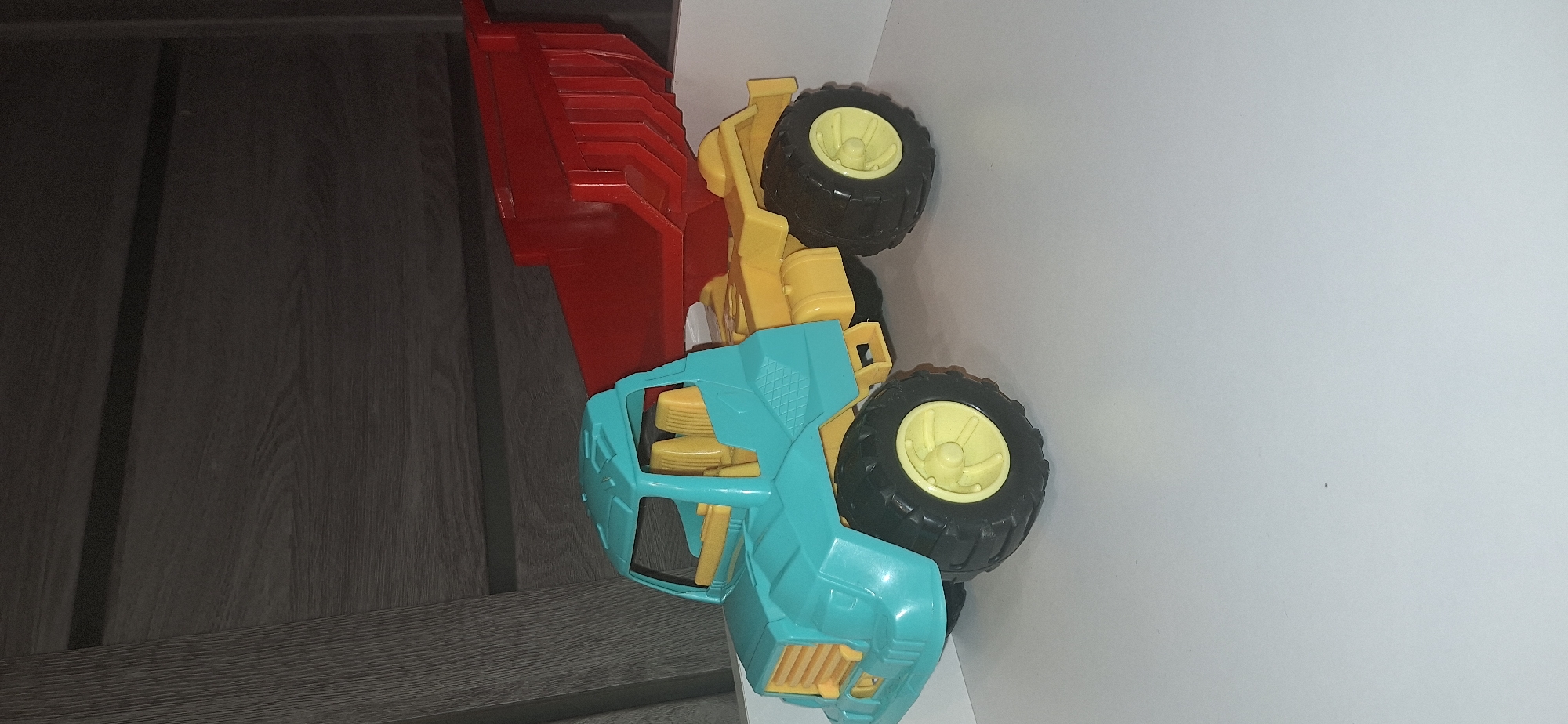 A toy dump truck is shown in the center of the image. The dump truck is primarily teal and yellow, with a red dump bed. The truck has four large black wheels with yellow rims and prominent treads. The cab of the truck is teal with yellow seats visible inside. The dump bed is raised and is red with a ribbed texture. The truck is positioned on a white surface, possibly a table or shelf. Behind the truck is a dark, textured background that appears to be a wooden surface, possibly a wall or furniture. The lighting in the image is somewhat dim, casting shadows from the truck onto the white surface. The image does not contain any people or animals. The location of this scene in Bila Tserkva, Ukraine cannot be determined from the image.