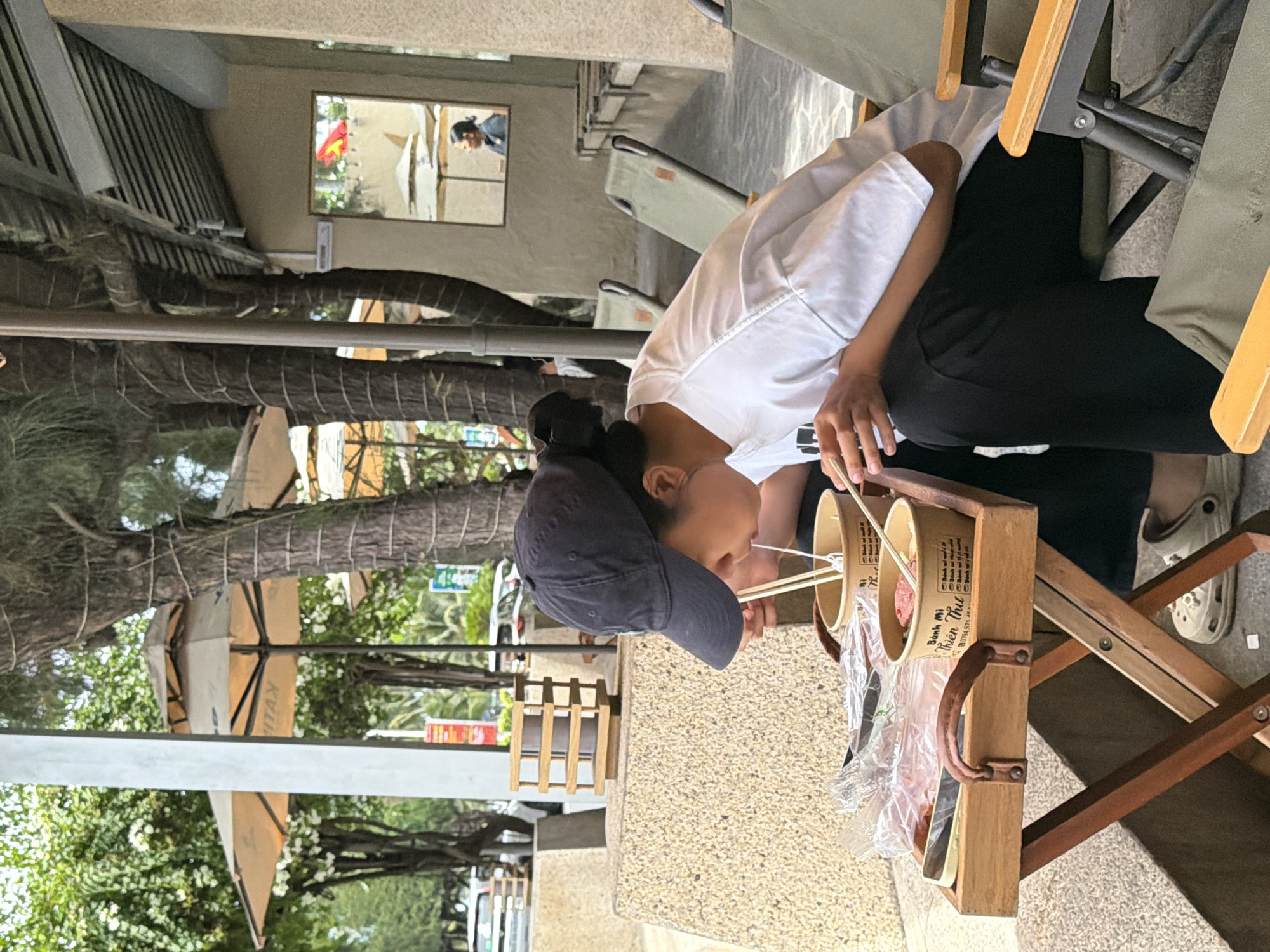 A young person wearing a black baseball cap and a white t-shirt is eating noodles with chopsticks from a disposable bowl. The person is seated at a small wooden table outdoors, next to a person in a reflection in a mirror. The table is positioned on a paved area in front of a building with a beige facade. In the background, trees with rough bark and large umbrellas provide shade. Further back, cars and a street are visible. The scene appears to be a casual dining setting in Nha Trang, Vietnam, likely a street-side cafe or restaurant, given the urban environment and the presence of outdoor seating.