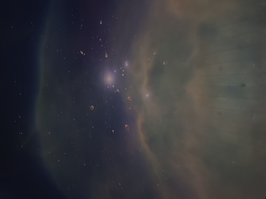 The image displays an interior view of a dark, domed projection space, likely a planetarium, located in Canela, Brazil. The environment is dimly lit, illuminated solely by the projected imagery. The central subject is a projection of a cosmic scene featuring a deep blue to purple background with numerous white and faint blue point light sources representing stars. Swirling, diffuse formations in shades of muted green, brown, and light orange, indicative of nebulae, are prominent across the mid-section. Bright, concentrated light clusters are visible within the projection. The lower portion of the image reveals the curved, paneled surface of the projection dome, characterized by faint vertical lines indicating structural seams. Scattered across the entire projected image are numerous irregularly shaped, opaque specks, varying in size, consistent with dust particles or condensation on a camera lens or the projection surface; some appear roundish, while others are elongated or tear-drop shaped. No individuals, specific objects beyond the projection and dome structure, or observable actions/interactions are present in the scene.