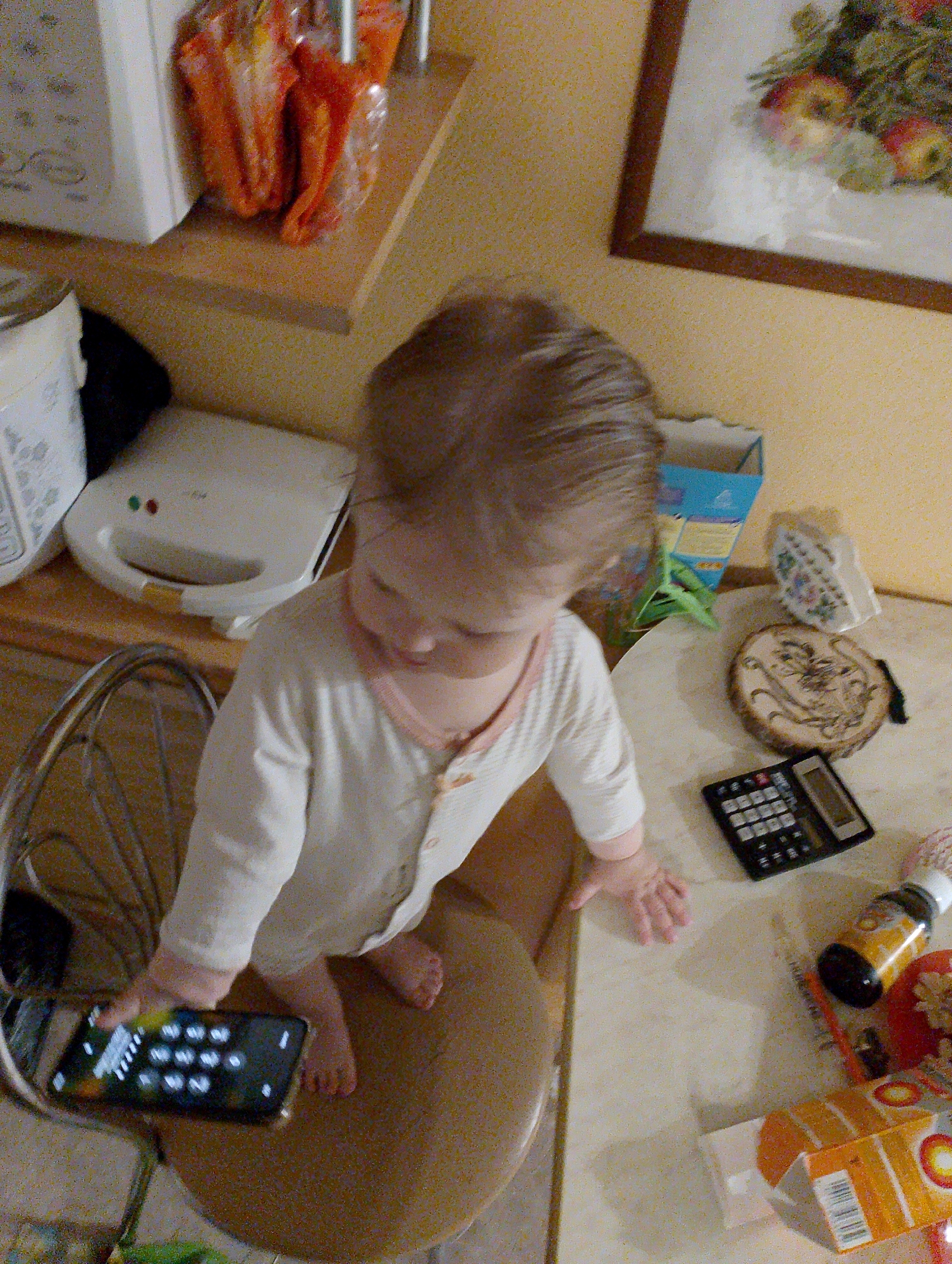 This image depicts an indoor scene in the city of Chaikovsky, Russia, interpreted as a residential kitchen or dining area. A young child, appearing to be a toddler with light brown hair, is positioned centrally, standing barefoot on a round brown stool. The child wears a light-colored, long-sleeved shirt with buttons and light pink trim on the collar and cuffs. In their left hand, the child holds a black smartphone displaying a numeric keypad. Their right hand rests on a light-colored countertop.
On the countertop, to the child's right, a black electronic calculator is present, alongside a round wooden object featuring a burned-in design. Further right on the countertop, a white ceramic item with a floral pattern, an open orange cardboard box, and a yellow-labeled bottle with a black cap are visible. In the background, to the left, a white sandwich maker with illuminated red and green indicator lights rests on a wooden surface. Above it, on a shelf, several orange plastic bags are stored. The background wall is a light yellowish-tan, featuring a framed picture depicting a still life with fruit and leaves.