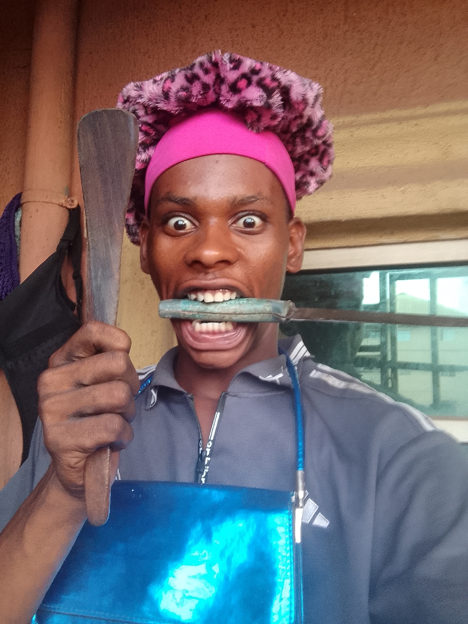A person is holding a large wooden spatula in their right hand and a metal object in their mouth. The person is wearing a pink and leopard-print head covering, a gray shirt with a blue lanyard and a blue apron. The background includes a window and a wall. The location is unspecified.