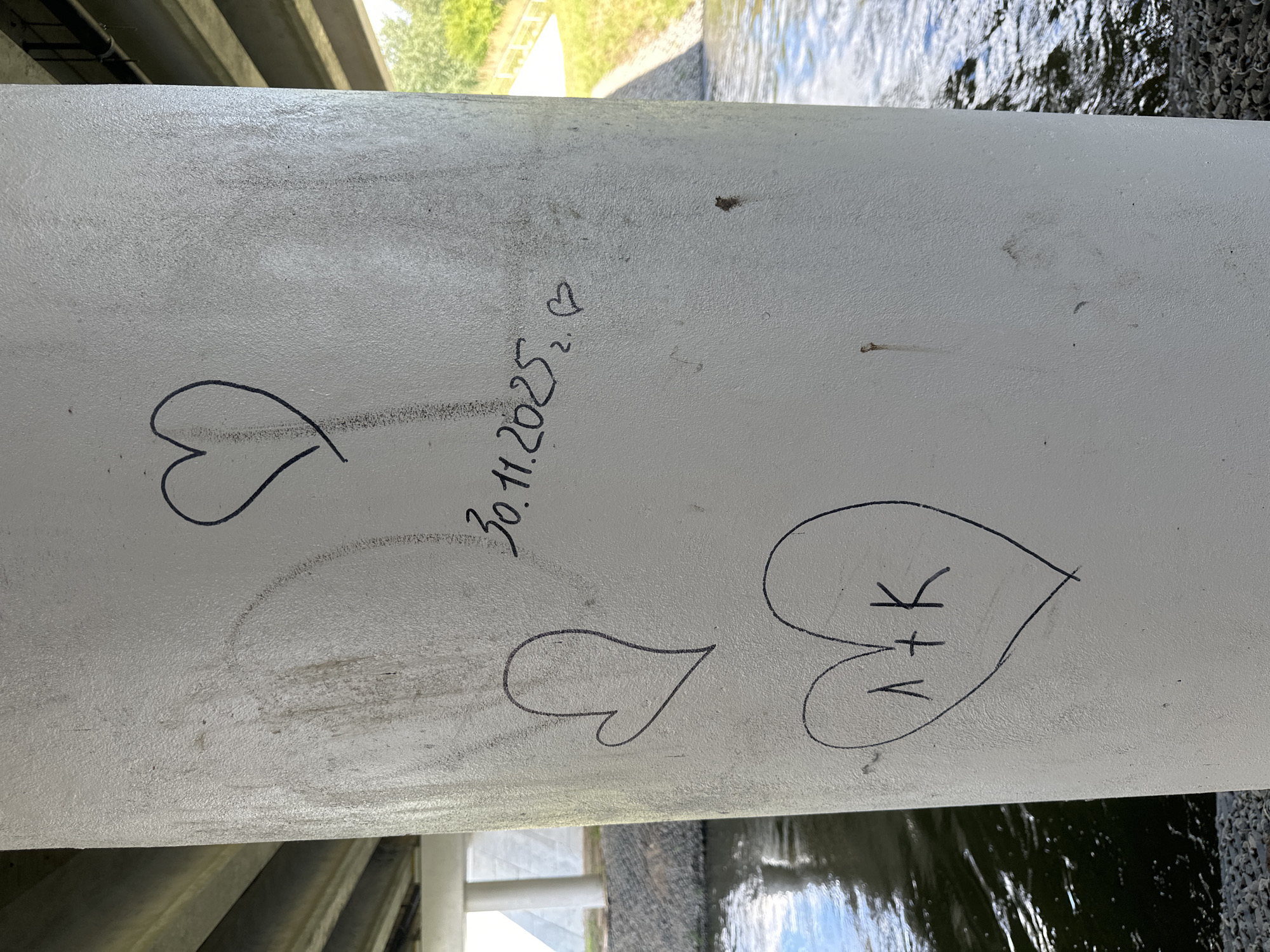 A concrete pillar supporting a bridge is covered in graffiti. In the upper portion of the pillar, a large, hand-drawn heart is visible, outlined in black marker. Below it, the date "30.11.2025" is written, followed by a smaller heart and the numeral "2". Further down, another heart shape encloses the inscription "1+K". To the left of the pillar, a body of water, likely a river, flows. The background shows trees and a grassy embankment. The pillar appears to be located in a park or recreational area within the city of Minsk, Belarus.