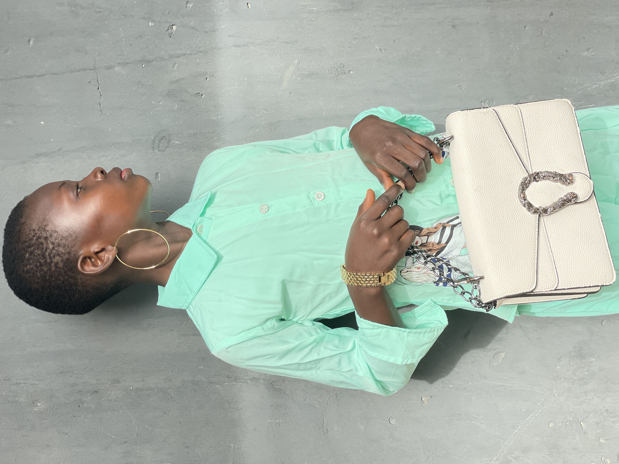 A Black woman with short hair lies on a gray surface. She wears a light green shirt dress, large gold hoop earrings, and a gold watch. She has a white purse with a silver clasp on her chest. The location is not identifiable as Badagry, Nigeria, based on the provided image.