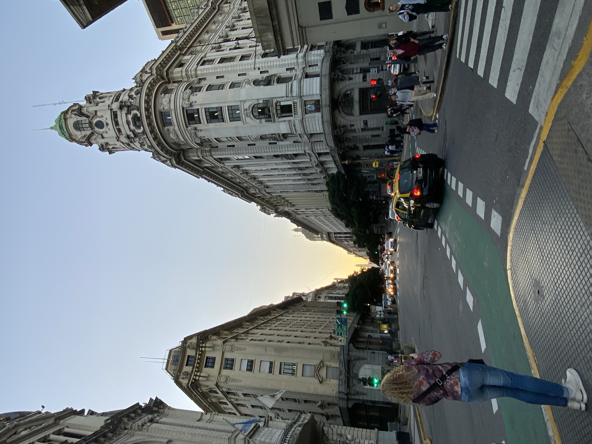 The image is a street-level shot in Buenos Aires, Argentina, featuring prominent architectural buildings. The street appears to be a major thoroughfare with vehicles, including taxis, and pedestrians. The buildings are tall with detailed facades and some have domes on top. There is a crosswalk visible, with people crossing the street. A woman is standing in the foreground, seemingly observing or photographing the scene. The sky is visible between the buildings, suggesting it is either early morning or late afternoon.