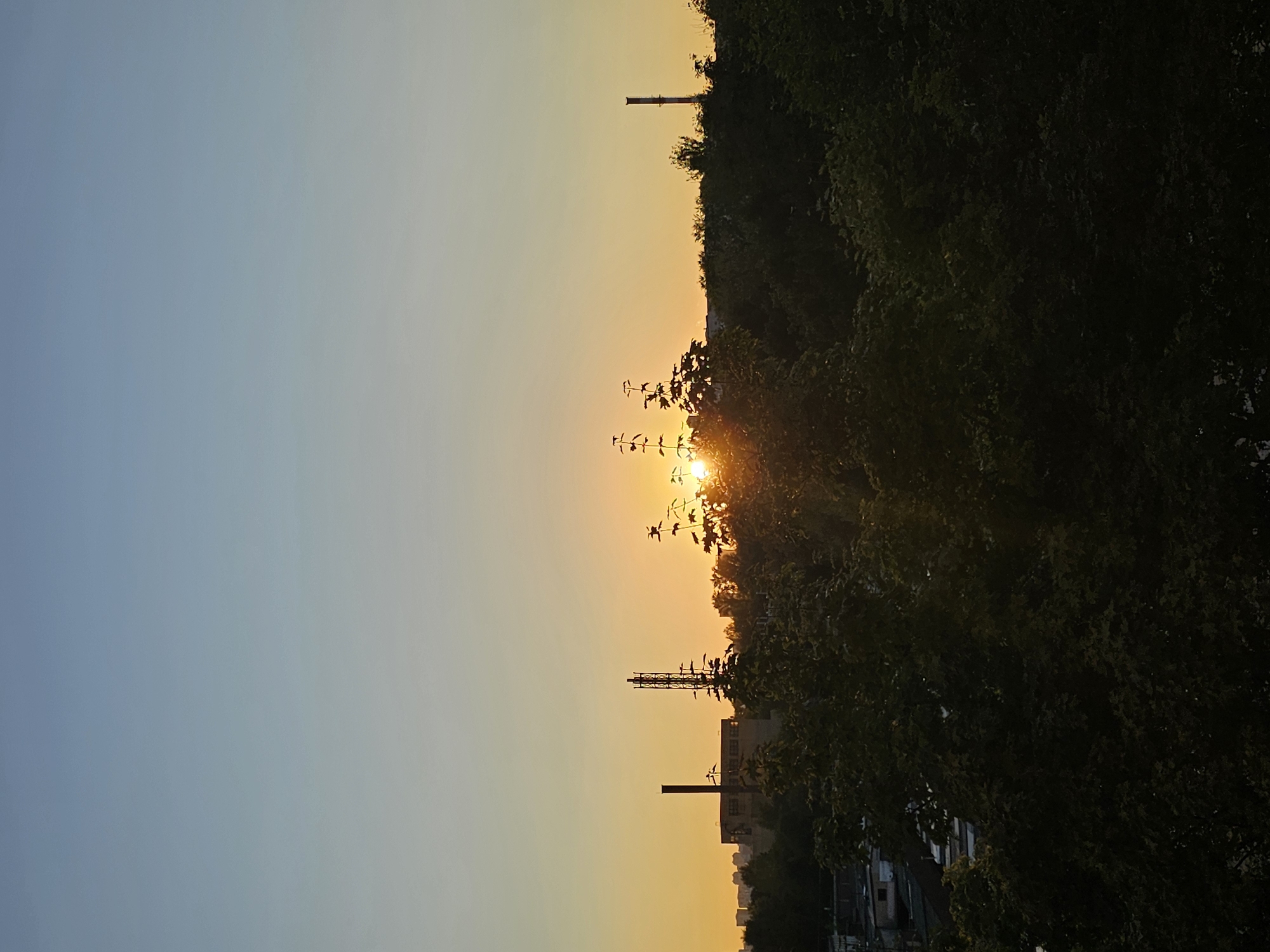 This is a landscape photograph taken at sunset over the city of Khimki, Russia. The sky is a gradient of light blue at the top, transitioning to a soft yellow-orange near the horizon, indicating the sun is setting. Silhouetted against this sky is a dense line of dark green trees in the foreground and midground. Beyond the trees, on the left side of the image, a tall, slender industrial chimney stands against the yellow sky. To its right, a lattice-like tower, possibly a communication tower, is visible, also in silhouette. Further to the right, another, shorter chimney is partially obscured by trees. In the far distance, the faint outlines of residential buildings can be seen. The sun itself is a bright, glowing orb peeking through the branches of the trees in the center of the frame, casting rays of light. The image is a still shot, capturing a moment of natural beauty combined with urban industrial elements. There are no people or animals visible in the frame.