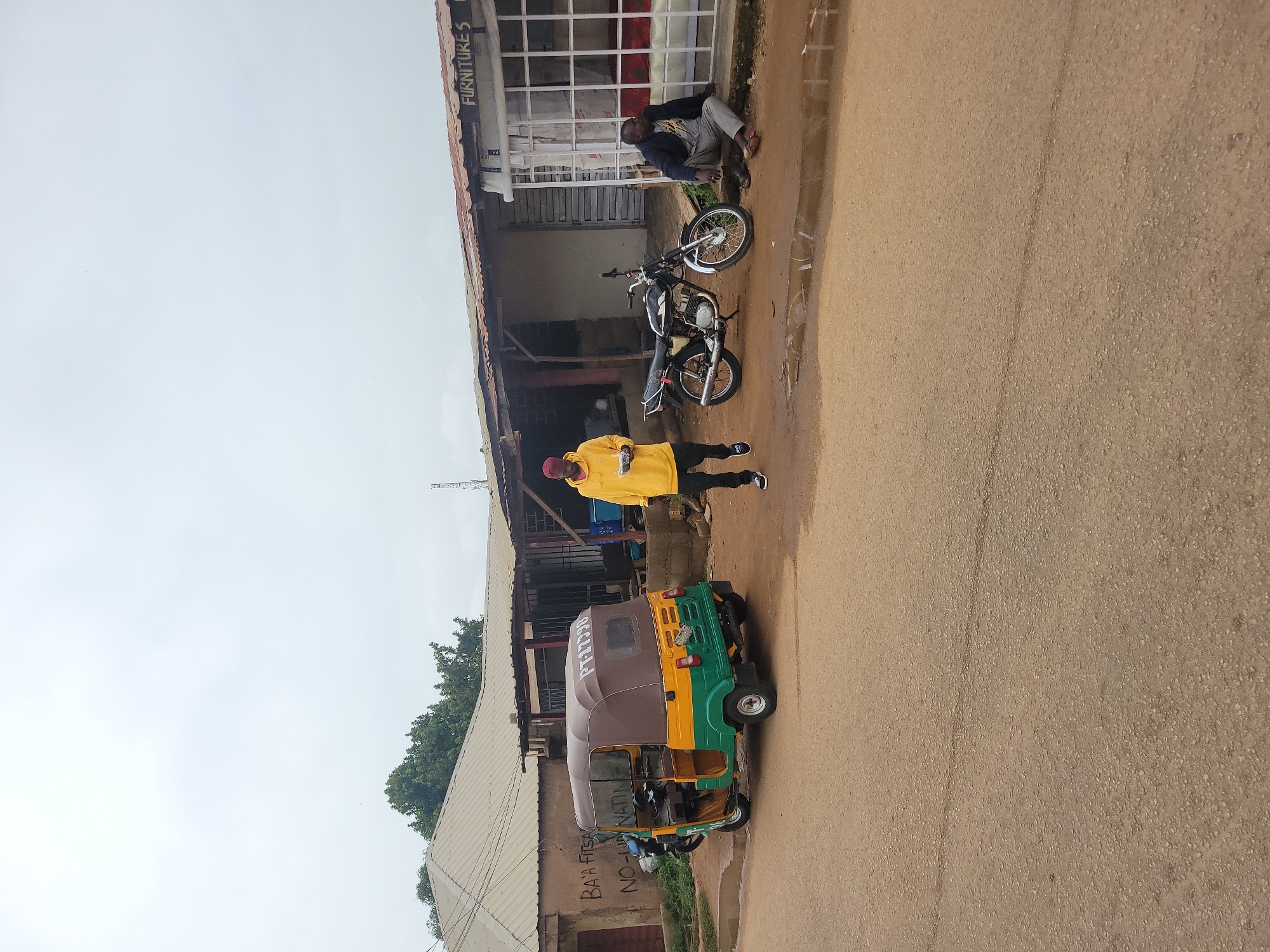 In Jos, Nigeria, on an overcast day, a street scene unfolds. A colorful auto-rickshaw, yellow, green, and brown, is parked on the right side of a paved road. To its left, a black motorcycle is parked. A man wearing a yellow hoodie and dark pants stands in the middle of the road, holding a white container. Further to the right, a man in a dark jacket and grey pants sits on a curb, facing the auto-rickshaw and motorcycle. Behind them, shops line the street with corrugated metal roofs. One shop has "FURNITURES S" visible on its facade. The ground is wet, with puddles reflecting the overcast sky, suggesting recent rain. The environment appears to be an urban setting with a mix of residential and commercial structures.