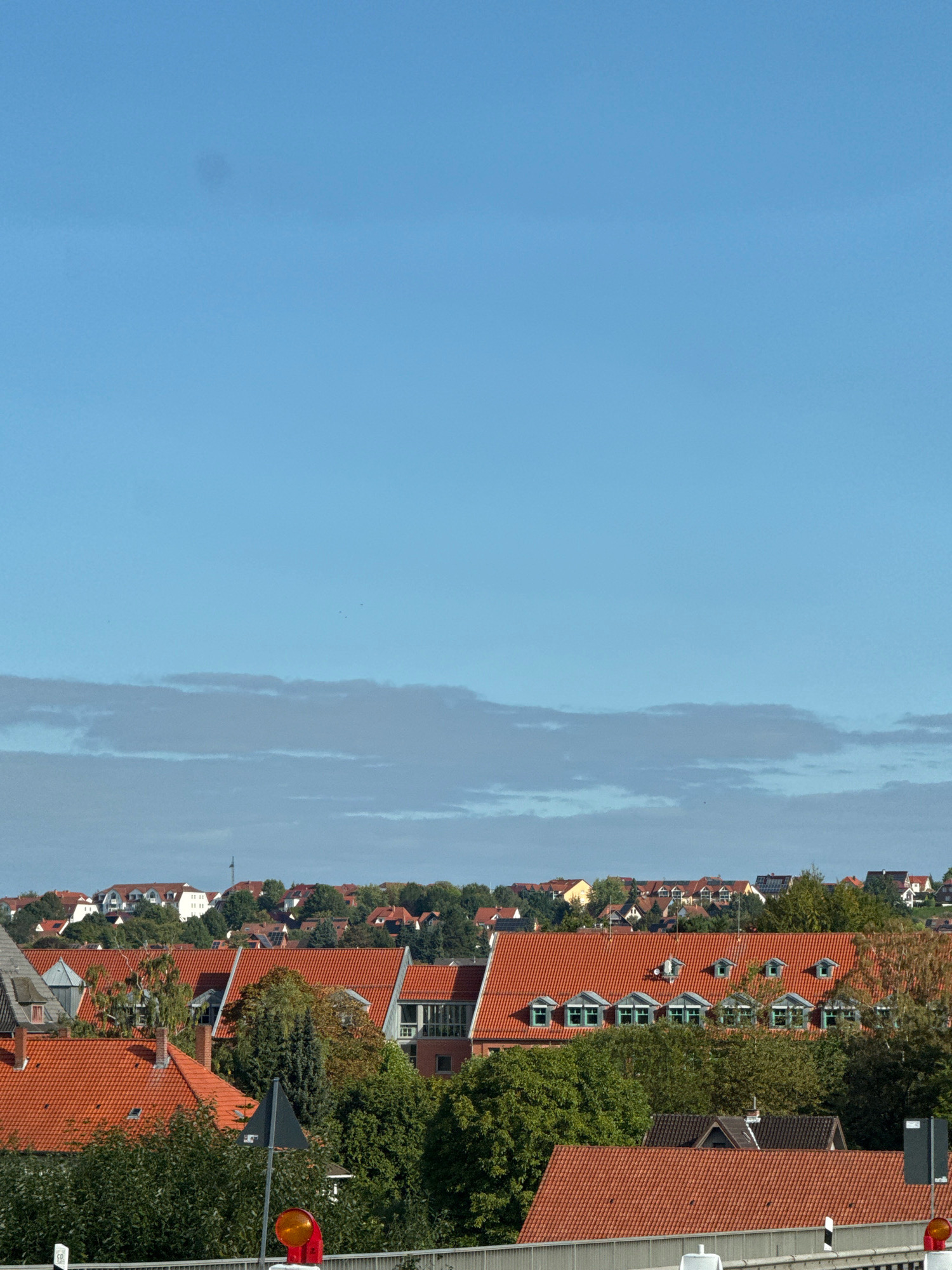 The image shows a panoramic view of the city of Osterode am Harz, Germany, with a clear blue sky above. In the foreground, there are several buildings with red tiled roofs, interspersed with lush green trees. A tall, dark gray traffic sign with a bright orange circular light is visible in the lower right portion of the image. In the background, more buildings with red roofs are nestled amongst the trees, covering the rolling hills of the city. The overall scene depicts a residential area with a mix of architecture and natural elements.