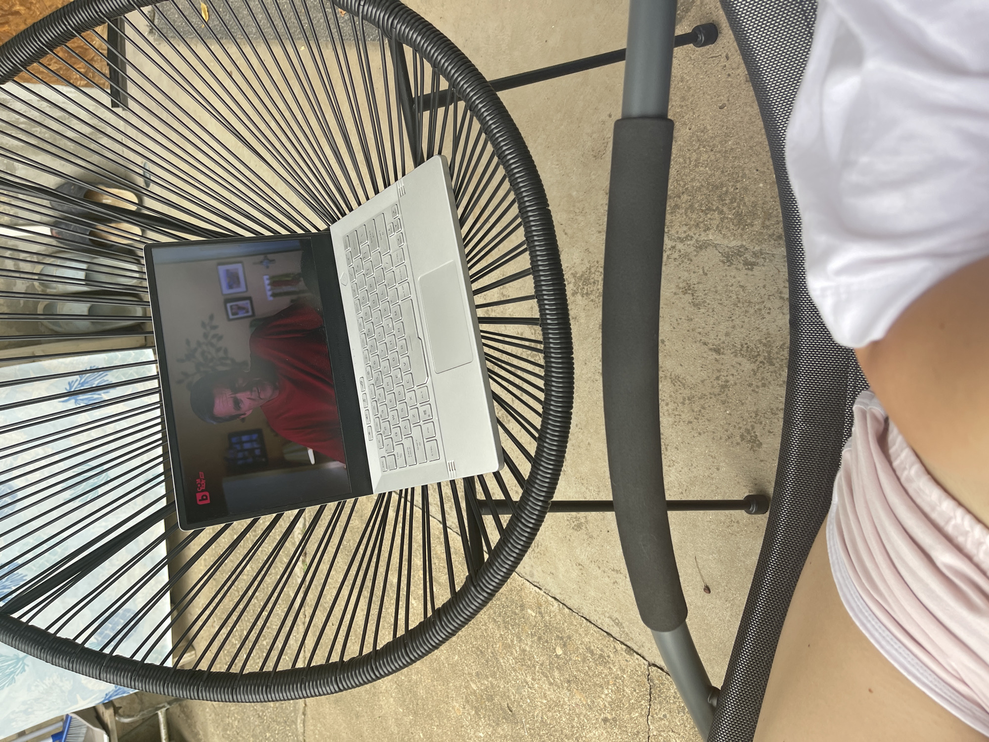 A top-down shot depicts a black, stringed outdoor table supporting a silver laptop with its screen displaying a video of a man in a red shirt. A portion of a gray metal chair with a padded armrest is also visible on the right. The environment suggests an outdoor setting, possibly a patio or backyard. The location in Sofia, Bulgaria, cannot be determined from the visual information provided.