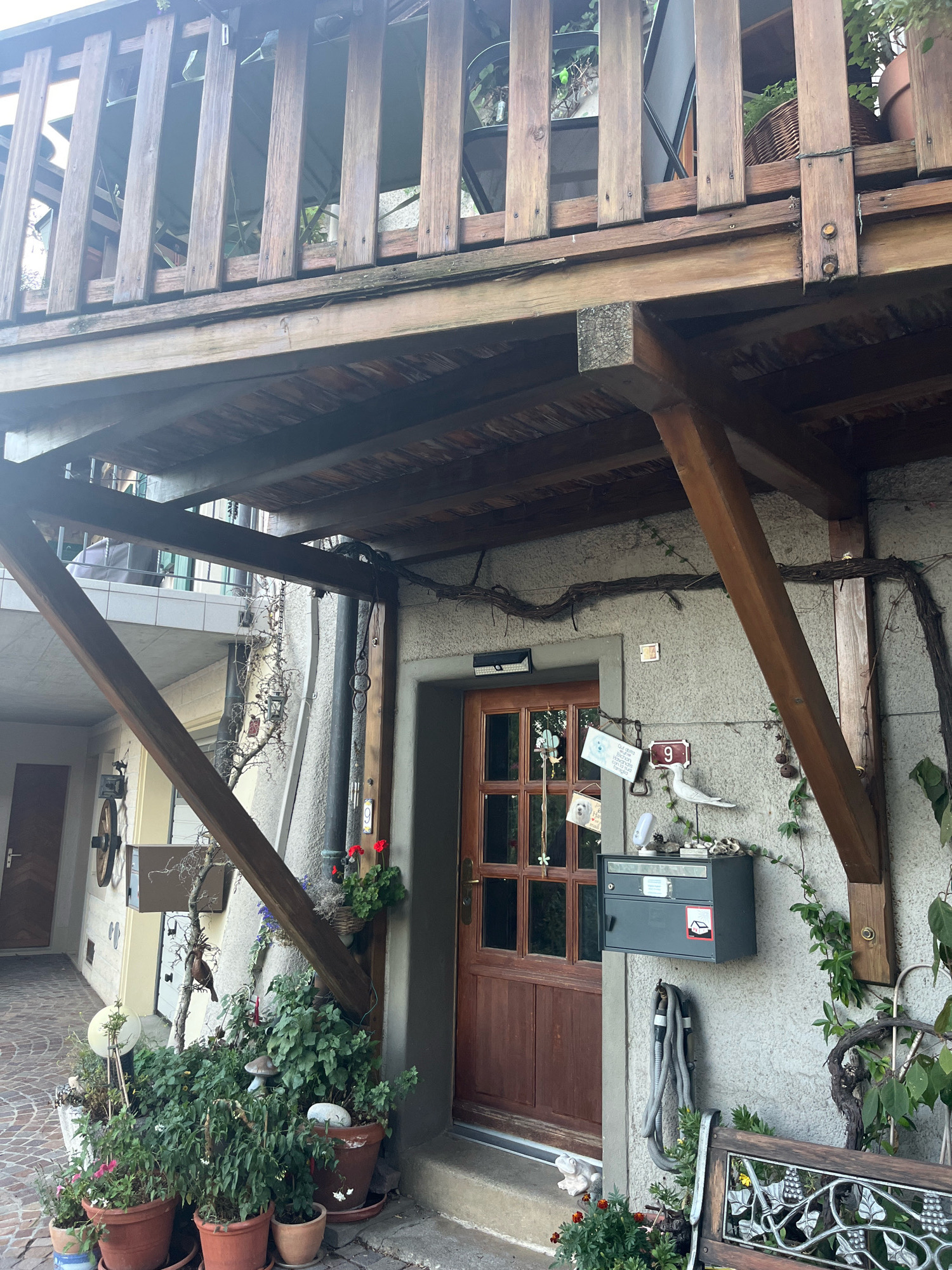 The image depicts the exterior of a building in Féchy, Switzerland, showcasing a rustic and green facade. A wooden balcony with vertical slats overlooks a lower level with a brown wooden door. The wall is adorned with decorative elements, including a grey mailbox, a white ceramic bird, and a sign with the number '9'. Various potted plants, featuring green foliage and some red and pink flowers, are arranged around the entrance, some on the ground and others hanging. A coiled garden hose hangs on the wall. The ground is paved with cobblestones. A section of a wrought-iron bench with a grape motif is visible in the foreground. The architecture suggests a charming, possibly older, residential property.