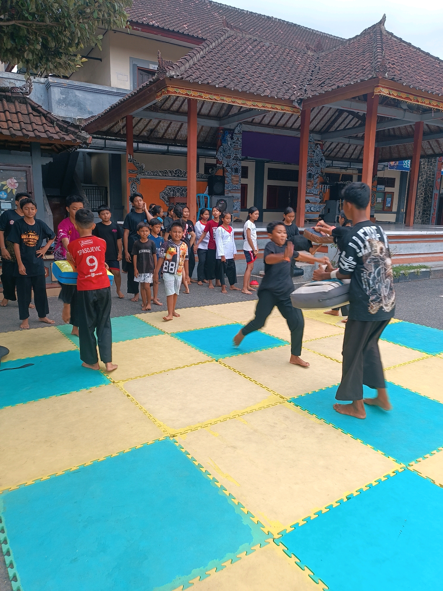 In Selemadeg, Indonesia, a group of young people are engaged in martial arts training on a mat made of interlocking puzzle pieces in blue and yellow. The setting appears to be an outdoor area adjacent to a building with a traditional Balinese tiled roof, supported by wooden pillars. Several boys and girls are standing and observing, while two boys are actively sparring. One boy, wearing a red shirt with the name "SPASOJEVIC" and the number "9" on the back, is holding a padded target. The other boy, dressed in black, is in motion, presumably striking the target. The ground is paved with interlocking mats that form a checkered pattern. The overall atmosphere suggests a community event or training session taking place in a cultural setting.