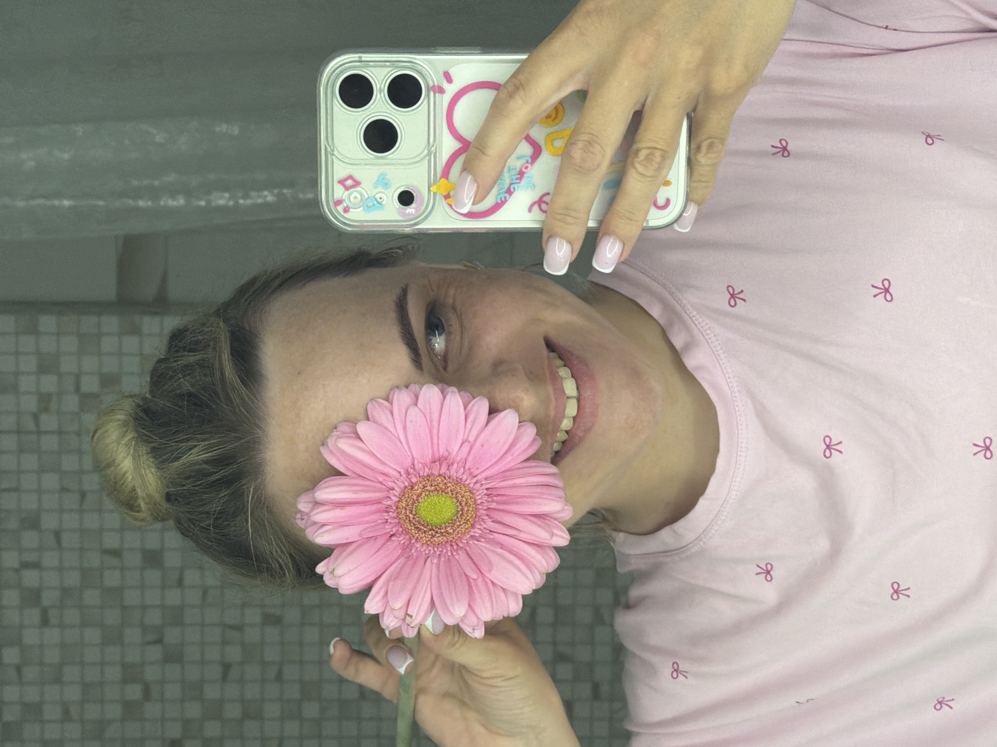 A woman in a light pink shirt with a bow pattern is holding a pink gerbera daisy in front of her face while taking a selfie with her smartphone. She is positioned with her head facing downward and slightly tilted. The phone, encased in a clear case with a cartoon design, is held by her right hand, which has manicured nails with a white tip. The background appears to be a tiled surface, possibly a bathroom or another area with similar tiling. There is no indication of this being specifically located in Votkinsk, Russia.