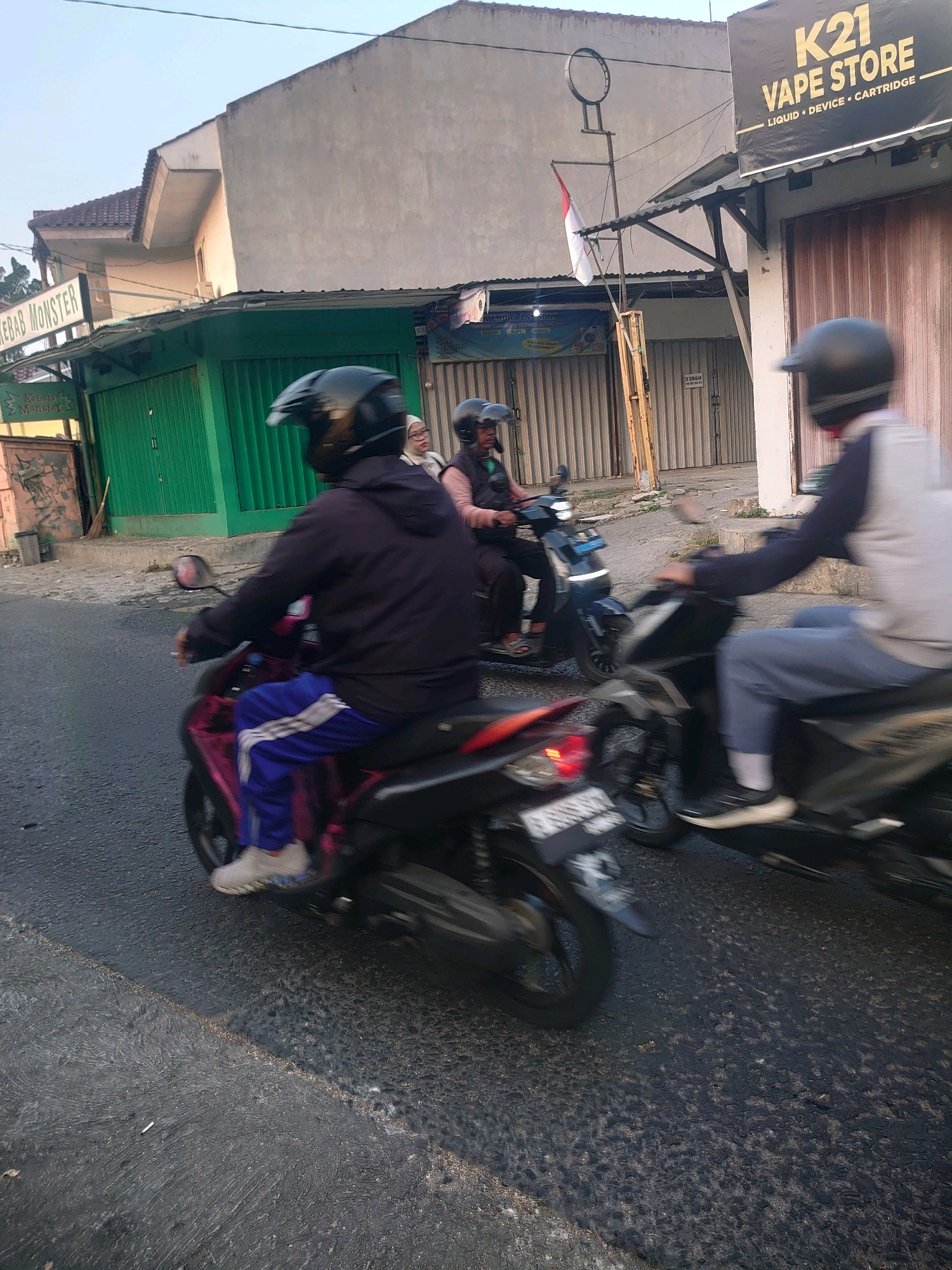 A medium shot captures three individuals riding motorcycles on a street in Depok, Indonesia. The foreground features a person on a pink motorcycle, wearing a black helmet and blue pants with white stripes. Their left leg is bent, resting on the footrest, and their right foot is on the ground. To the right, another person on a black motorcycle, wearing a gray and blue uniform and a black helmet, is in motion, with their body blurred. Further in the background, a third person on a dark motorcycle, wearing a black helmet, is also visible. The street appears to be asphalted, with some patches of worn texture. On the left side of the image, a building with a green shuttered storefront displays a sign that reads "KEBAB MONSTER". On the right, a building has a prominent black sign with yellow lettering announcing "K21 VAPE STORE" and "LIQUID • DEVICE • CARTRIDGE". Other shops with metal shutters are also present. An Indonesian flag is visible on a pole attached to a makeshift structure. The sky is overcast.