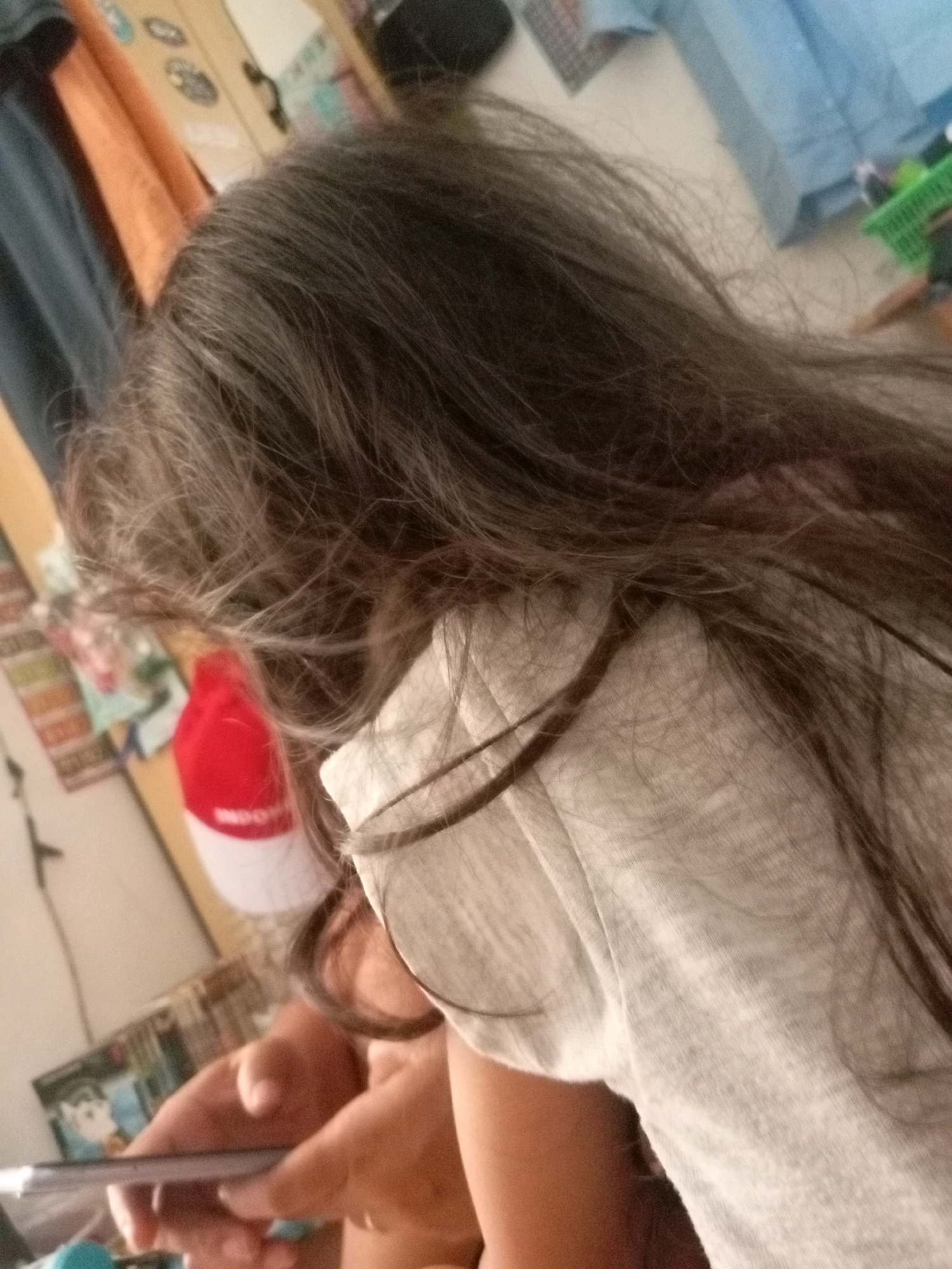 The image shows a person's head and shoulders, viewed from the side and slightly from behind. The person has long, dark, wavy hair that partially covers their face and shoulders. They are wearing a light gray hooded sweatshirt. In the background, there are various objects, including what appears to be clothing on a hanger, a red and white hat with writing on it, and some shelves or furniture with items on them. The overall environment suggests an indoor setting, possibly a bedroom or closet. The image is taken with a mobile phone and has a slight blur, particularly in the background. The location is identified as Batam, Indonesia.