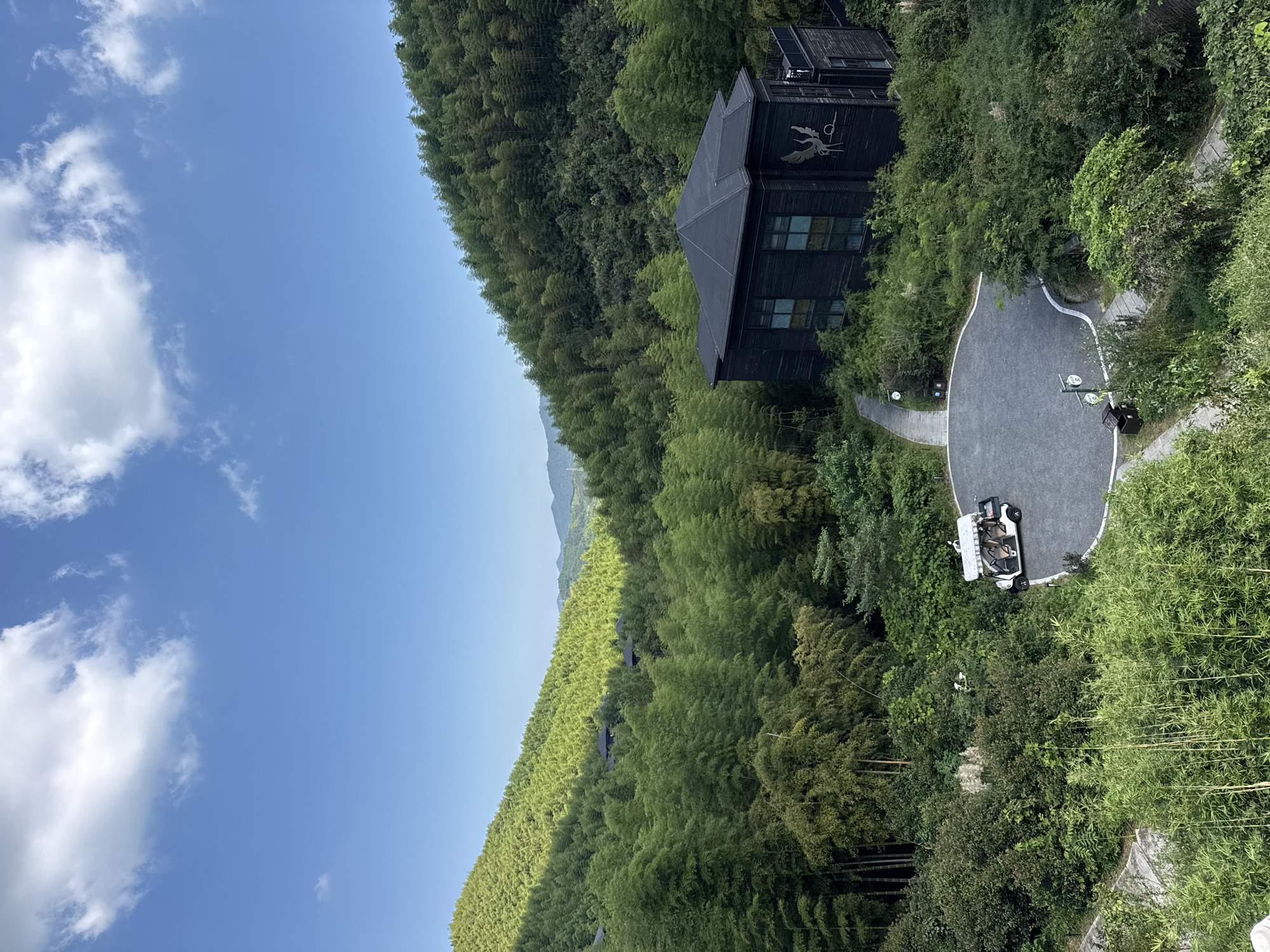 The image depicts a serene scene of a modern, dark-wood building nestled amidst a dense bamboo forest on a hillside. A white golf cart is parked on a gravel courtyard in front of the building. The sky is a clear blue with scattered white clouds. The architecture of the building is contemporary, with a dark, textured exterior and large windows. A metallic winged creature emblem is visible on the side of the building. The surrounding environment is lush and green, dominated by towering bamboo stalks that cover the rolling hills. Wuxing District in China is known for its beautiful natural landscapes, and this image captures that essence.