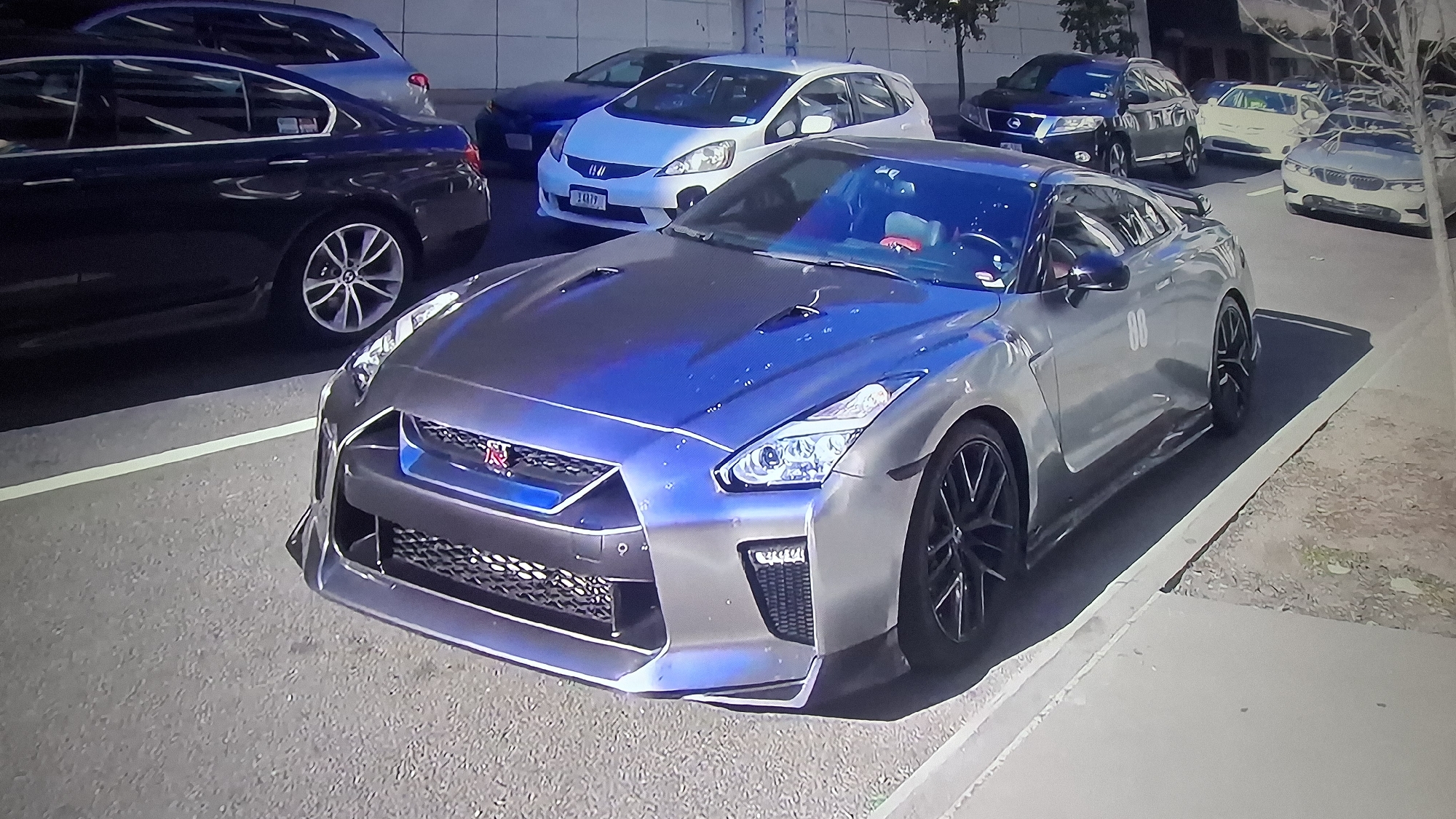 The image shows a silver Nissan GT-R parked on a city street. Other cars, including a white Honda Fit, a black BMW sedan, and a dark SUV, are parked nearby. The GT-R has black wheels and a prominent front air dam. The hood of the GT-R has a blue sheen. The number "88" is visible on the car's side. The setting appears to be an urban environment, possibly in an area with a mix of residential and commercial buildings. The presence of multiple cars suggests a relatively dense urban area. There are no visible people in the frame.