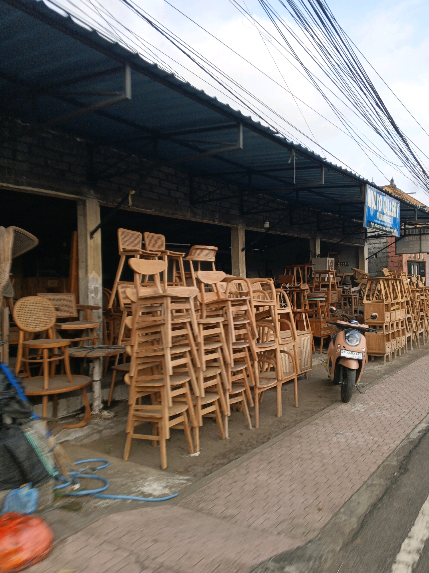 The image shows a street scene in Sukawati, Indonesia, in front of a furniture shop. The shop's facade features an awning supported by metal beams, and the building itself is constructed of dark brick. Numerous wooden chairs of various designs are stacked and displayed outside the shop, some with woven seats and backs. To the right, a light pink scooter is parked on a paved sidewalk. The sidewalk is made of red interlocking pavers and transitions to a gray concrete curb bordering a paved road. In the foreground, to the left, a dark blue hose is coiled on the ground, near a broom and a red, circular object. Overhead, a dense network of electrical wires crisscrosses the sky, which is partly cloudy. A blue sign with white lettering, reading "MULYO GALLERY FURNITURE", is affixed to the shop's exterior.