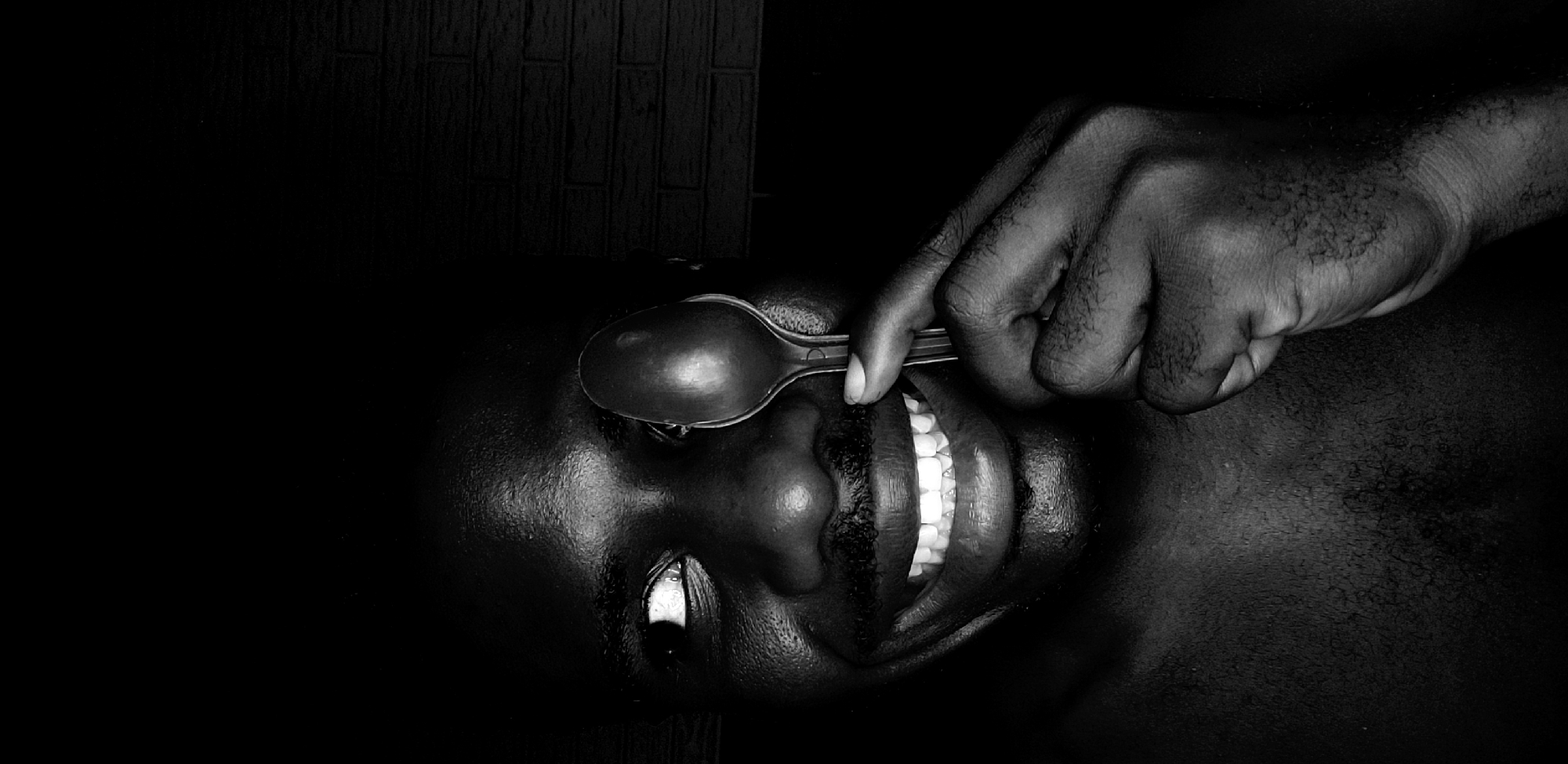 This high-contrast, monochromatic photograph presents a close-up, angled view of a person, likely an adult male, in a dark setting. The central subjects are the person's face, a hand, and a spoon.
The person's face is oriented sideways, with their left side towards the top of the frame. A dark-skinned hand holds a metallic spoon, pressing it onto or resting it over the person's left eyelid. The person's mouth is wide open in a pronounced smile, revealing bright white teeth, which stand out sharply against the dark skin. The visible right eye is open, looking towards the bottom of the frame, and appears glossy. Skin texture, including fine hairs on the hand and arm, is visible due to the lighting. The background is largely dark, with a faint, subtle brick-like pattern discernible in the upper section, suggesting an indoor or contained space. The lighting creates strong shadows and highlights, emphasizing contours and textures.
The image depicts an unusual and potentially unsettling act, juxtaposing a broad smile with the spoon pressing against the eye, creating an ambiguous emotional tone that could suggest discomfort, performance, or surrealism. There is no visible text in the image. The scene is dark, implying night or a poorly lit environment.