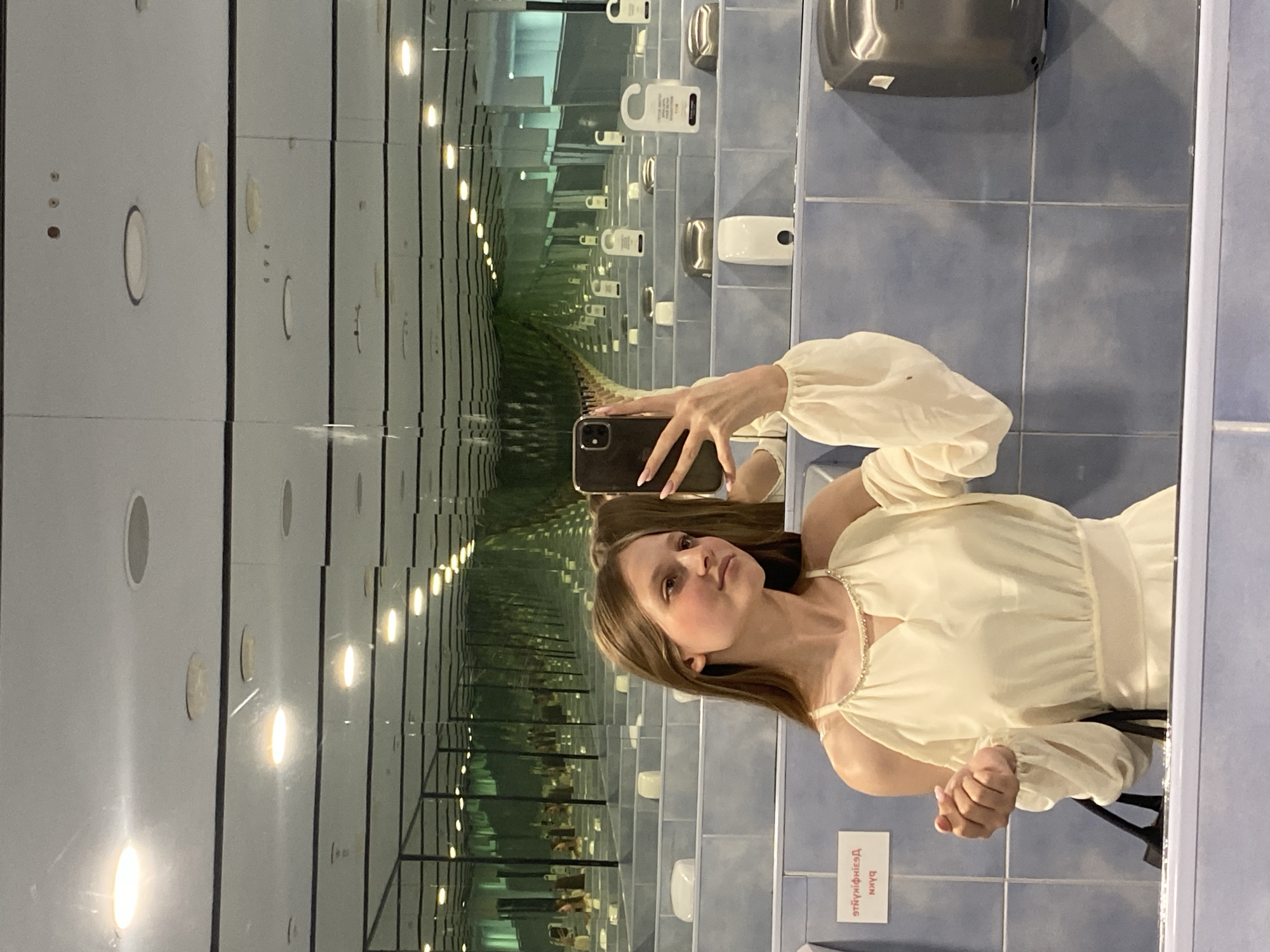A young female with long, straight, light brown hair, light complexion, and minimal visible makeup takes a mirror selfie in what appears to be a public washroom or restroom. She wears a cream or off-white cold-shoulder dress with a V-neckline adorned with a pearl or beaded trim, and long sleeves with elasticated cuffs. She holds a dark-cased smartphone with a triple camera array in her right hand, positioned to capture her reflection. A strap from a black bag is visible in her left hand.
The environment creates an "infinity room" illusion due to extensive mirrored surfaces on the walls and possibly the ceiling, reflecting fixtures into an infinitely receding corridor. The lower half of the walls is tiled with light blue/gray rectangular tiles, separated by dark grout lines. Reflected along the mirrored corridor are multiple white ceramic sinks or toilet basins, separated by blue tiled partitions. Above the sinks, various hygiene dispensers are visible, including a silver hand dryer on the right and a white soap dispenser. White rectangular signs with black text, displaying Cyrillic script in reverse ("ЯТНУЯІНІЧІЕЯД" and "NXYq" are discernible on the left side) are affixed to the tiled wall. Further back in the reflections, several white "Do Not Disturb" style door hangers with text and icons are seen. The white ceiling features numerous evenly spaced, recessed circular light fixtures, casting bright, warm illumination that is also amplified by the mirrors. The floor is not visible.
The specific city and country are Unknown, but the presence of Cyrillic script on the signage suggests a location within a region where this alphabet is used.