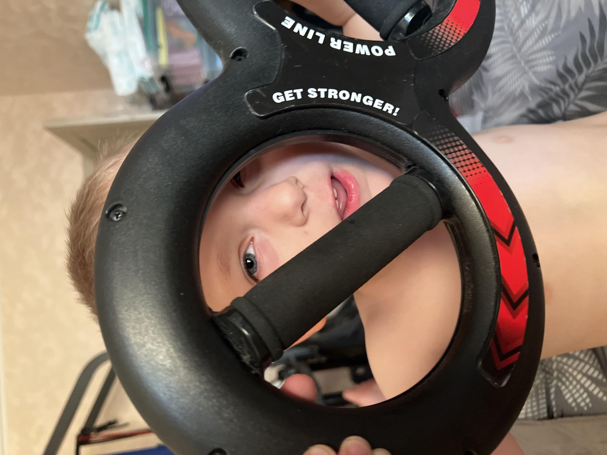 A child is looking through the center of a black, figure-eight-shaped exercise device with handles. The device has "POWER LINE" and "GET STRONGER!" printed on it. Red arrow designs are visible on the device. The child has fair skin, blond hair, blue eyes, and a slightly open mouth. A patterned shirt and a shelf are visible in the background. There is no indication of this image being in the city of Mytishchi, Russia.
