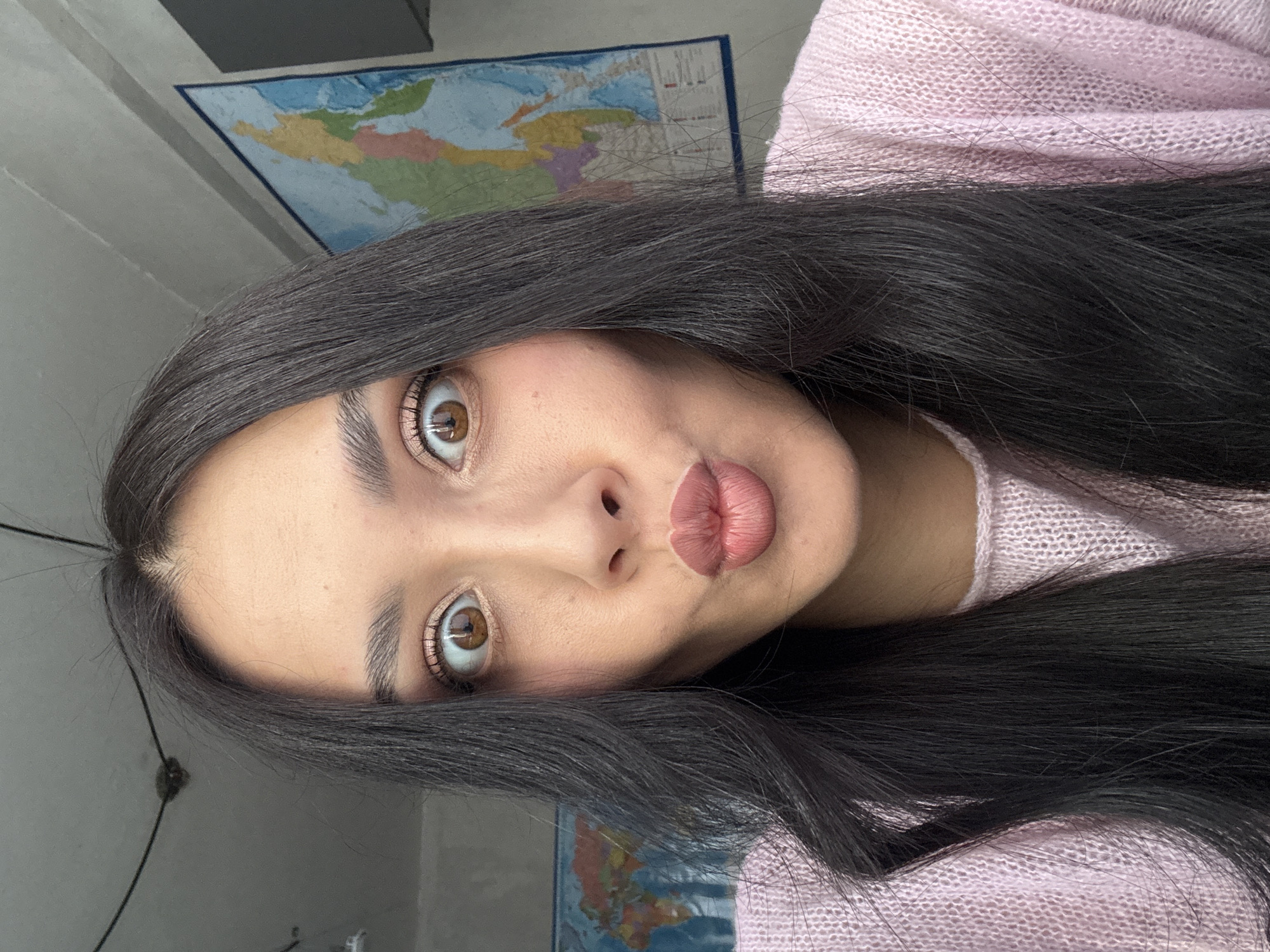 A young woman with long, dark hair is centered in the frame, wearing a light pink sweater. She is looking directly at the camera with a slight pout. Behind her, partially visible, is a world map. The background appears to be a room with neutral-colored walls. There is no indication of the city of Votkinsk, Russia, or any specific location.