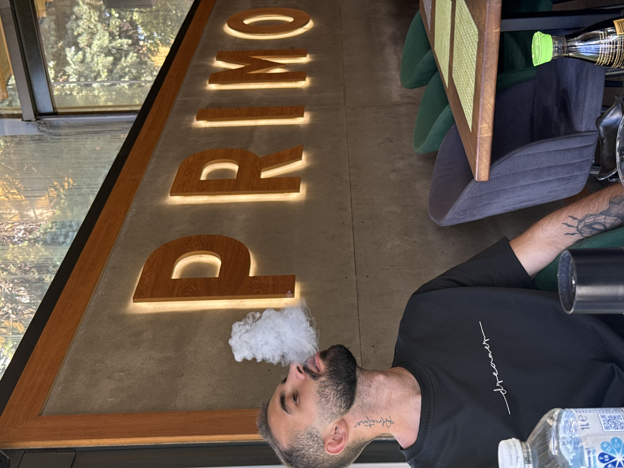 A man with a beard and tattoos is exhaling a cloud of smoke in front of a wall with illuminated letters that spell "PRIMO". He is wearing a black t-shirt with the word "dreamer" written on it in white script. In the foreground, a plastic bottle of water and a dark cylindrical object are visible. Behind the man, there is a table with green placemats and chairs. The environment appears to be an indoor seating area, possibly a restaurant. The exterior view through a window shows trees. The man is positioned to the left of the frame, with the "PRIMO" sign extending across the background.