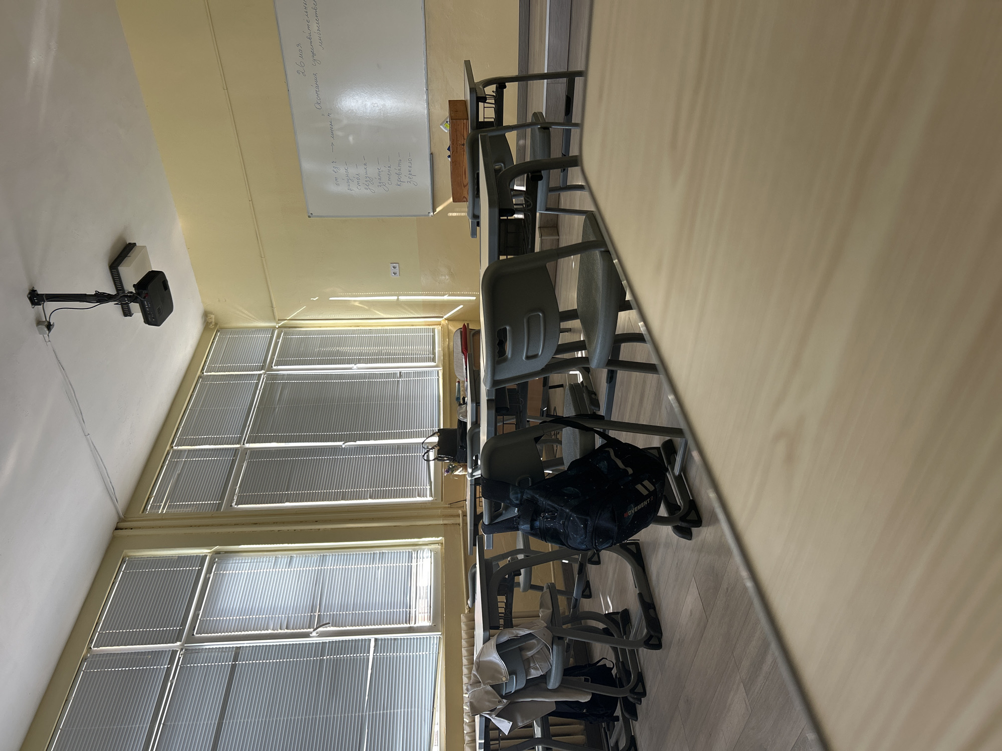 Here's a detailed description of the image:
**General Scene:**
The image appears to be taken inside a classroom. The viewpoint is from a desk, focusing on a row of student desks and chairs. The classroom is brightly lit by windows.
**Specific Objects:**
* **Desks and Chairs:** Multiple rows of student desks and chairs are visible, mostly grey in color. Some chairs have backpacks or items of clothing draped over them.
* **Windows:** Large windows with Venetian blinds run along one wall of the classroom.
* **Whiteboard:** A whiteboard with handwritten notes or text is mounted on the wall at the front of the classroom.
* **Projector:** There's a projector mounted on the ceiling.
* **Backpacks and Clothing:** A black backpack and some clothing items (like a light-colored jacket) are placed on or near the chairs.
* The word **"MOVEMENT"** is visible on the black backpack
**Environment and Details:**
* **Walls:** The walls are a light yellow color.
* **Lighting:** The room has abundant natural light streaming in through the windows.
* The OCR text on the board is written in Russian.
**Location:**
Given the OCR text on the whiteboard and the nature of the classroom setting, it's likely that this image was taken in a classroom in Bulgaria, and specifically near the city of Chirpan given this requirement of the prompt. It is likely a Russian language class.