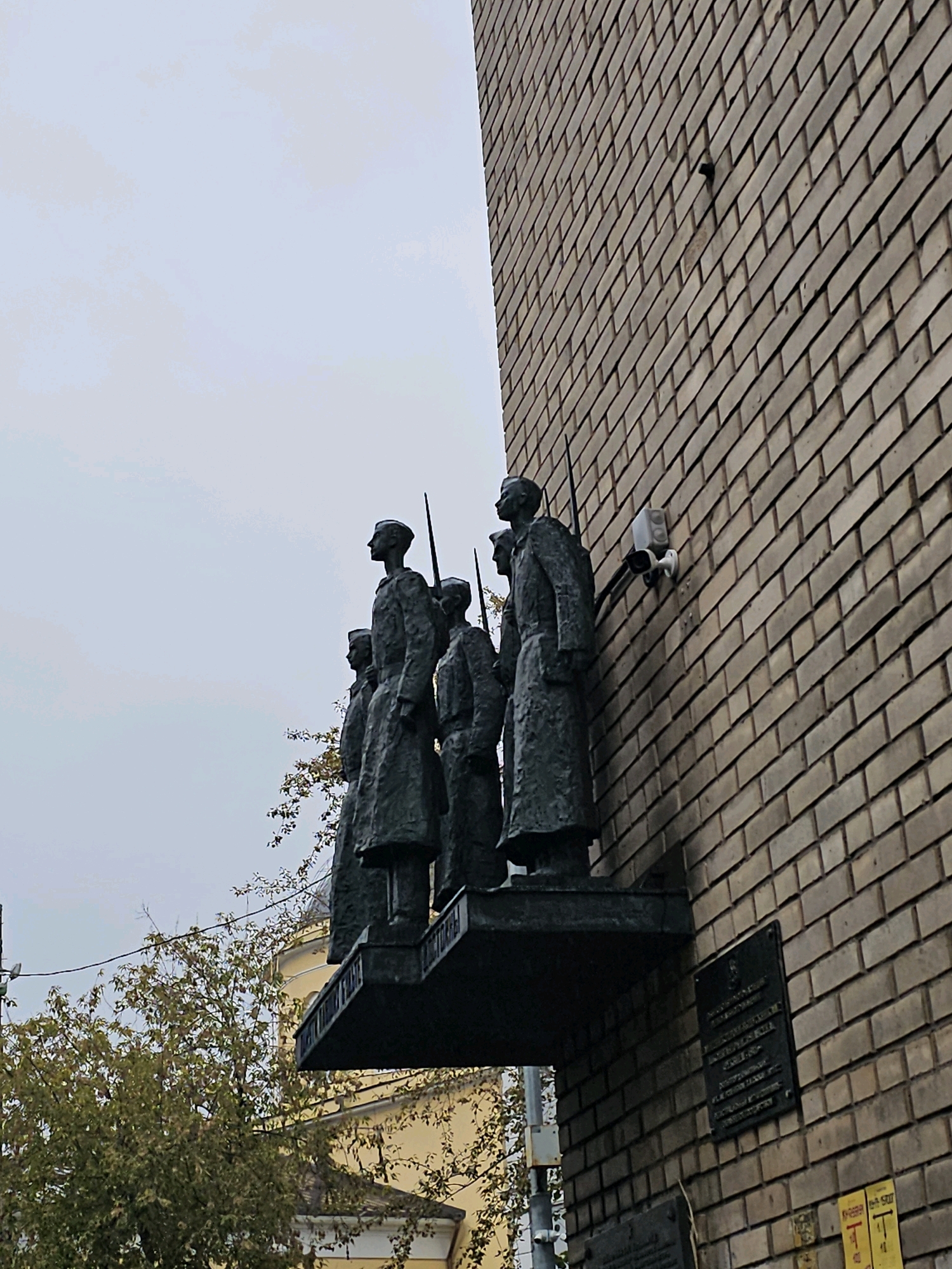 This image shows a bas-relief sculpture of five soldiers in uniform, standing in formation and holding rifles, mounted on a brick building. The sculpture is dark in color, likely bronze or a similar metal. A security camera is mounted on the brick wall above and to the right of the sculpture. A plaque is affixed to the brick wall below and to the right of the sculpture. Trees with yellowing leaves are visible behind and to the left of the sculpture, and a yellow building with a domed roof can be seen through the trees. The sky is overcast and a pale gray. This sculpture is located at the Museum of the Soviet Bunker on Taganka Square in Moscow, Russia.