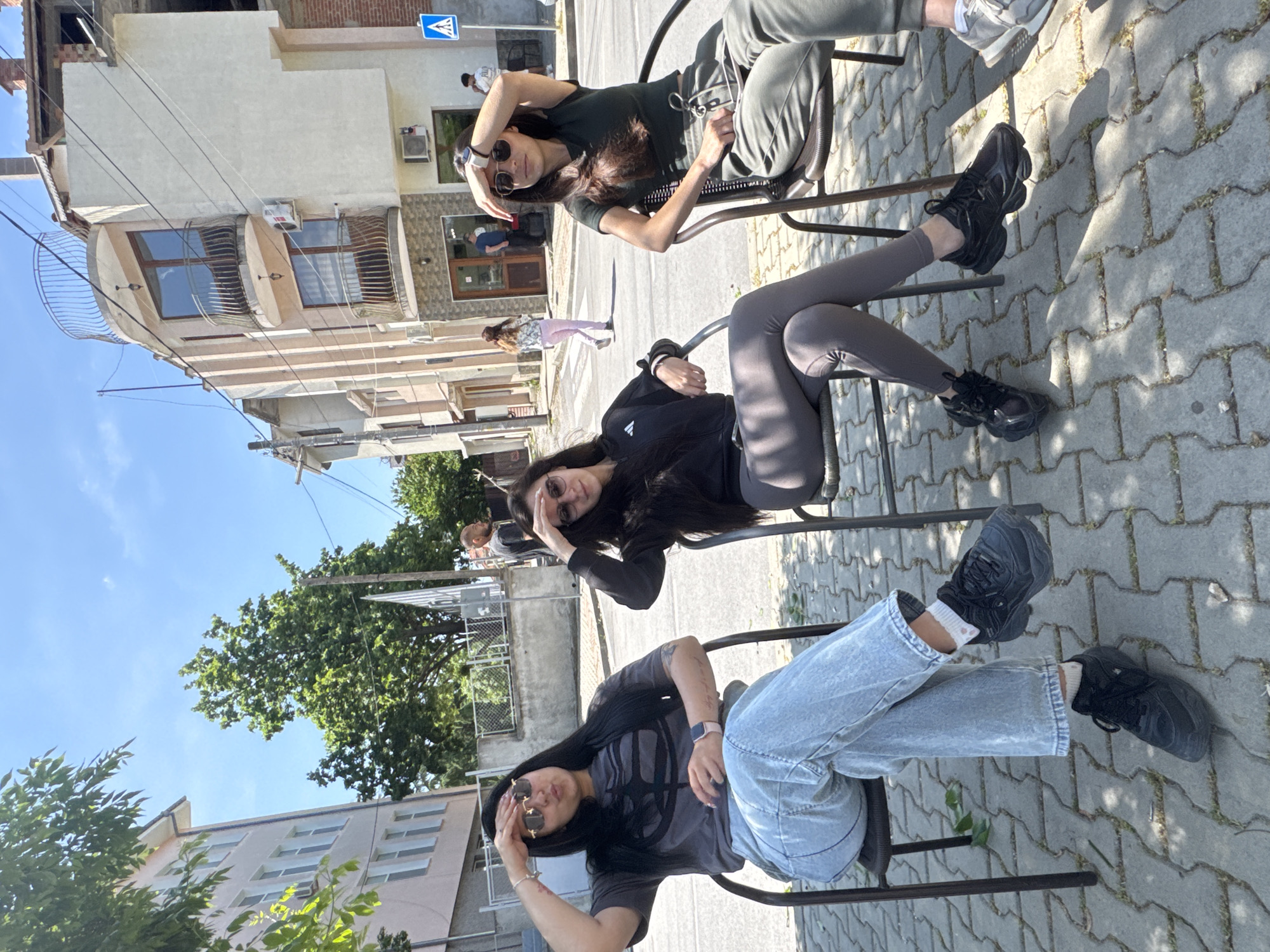 Three young women are sitting on metal chairs on a cobblestone street, outdoors in the city of Unknown, Unknown. The woman in the foreground is wearing light blue jeans and a dark t-shirt, and sunglasses. The woman in the middle is wearing dark leggings and a black sweater. The woman in the background is wearing dark green pants and a dark shirt. All three women are wearing sunglasses. The street is lined with buildings, and there are trees in the background.